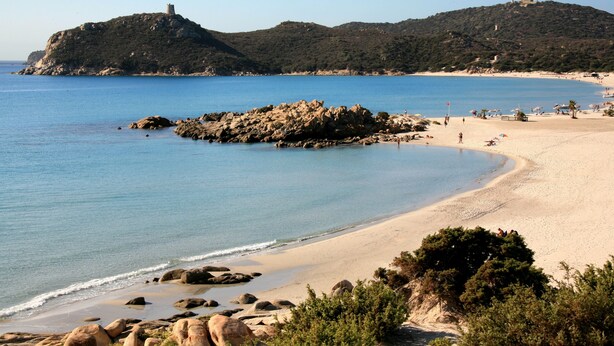 A coastal beach with rocky outcrops, clear blue water, and a distant hill with a tower. - Hotel Dell'Ancora (Villasimius)
