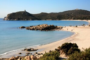 Una spiaggia costiera con affioramenti rocciosi, acque blu cristalline e una collina lontana con una torre.