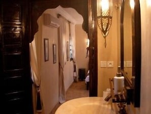 Un couloir étroit, avec une porte en bois sur la gauche, un miroir sur la droite et un petit lavabo, près duquel est accrochée une serviette.