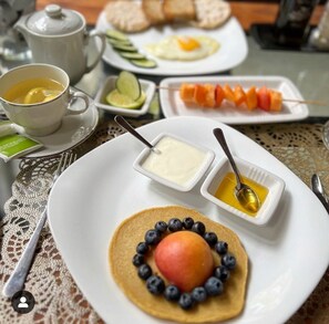 Desayuno con una pila de panqueques cubiertos de arándanos y fruta, dos recipientes pequeños de acompañamientos, y una taza de té.