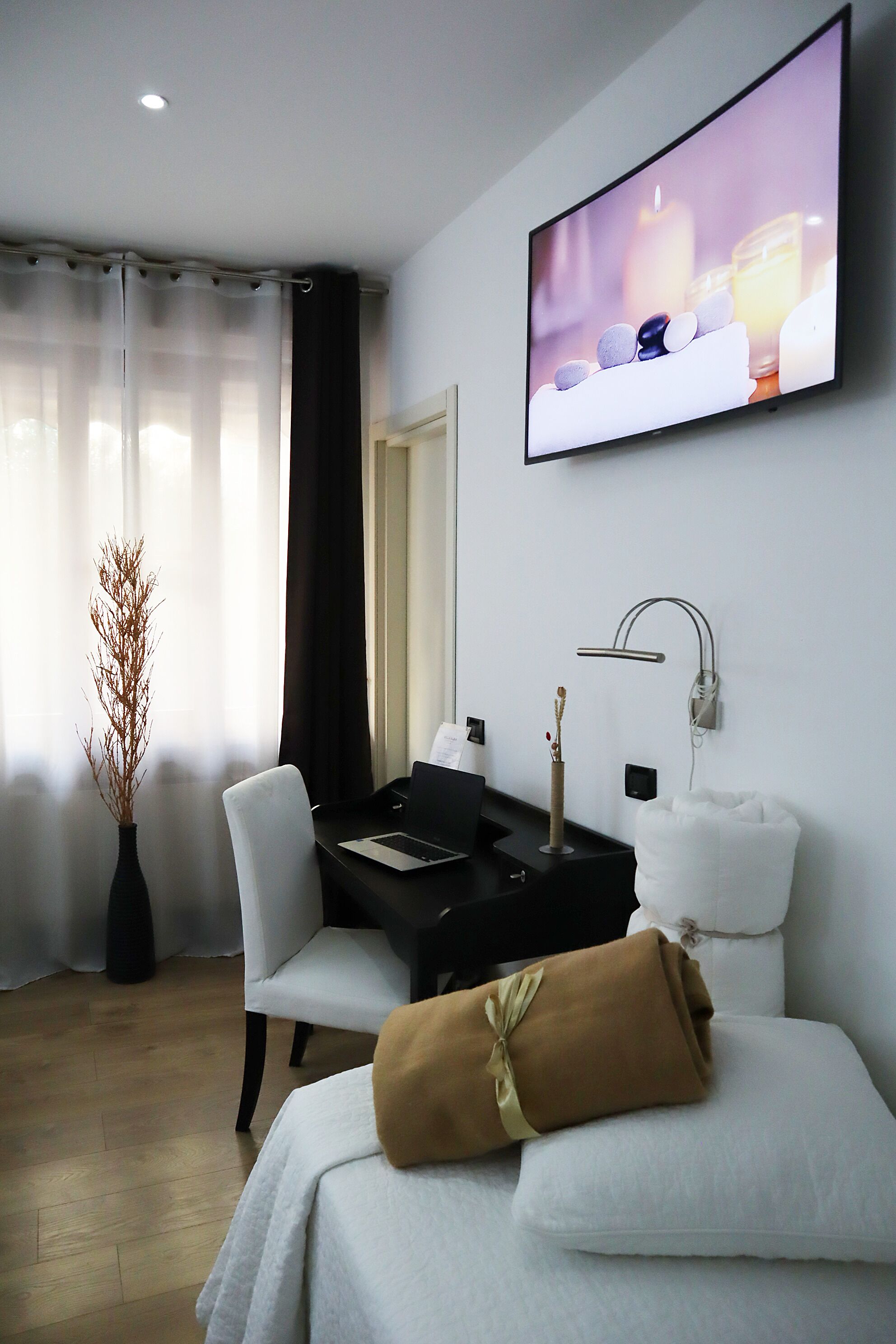 A modern hotel room with a flat-screen TV, a desk with a laptop, a vase with dried plants, a white chair, a bed with a brown towel, and a wall-mounted lamp.
