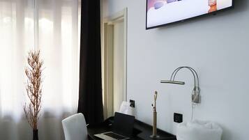 A modern hotel room with a flat-screen TV, a desk with a laptop, a vase with dried plants, a white chair, a bed with a brown towel, and a wall-mounted lamp.