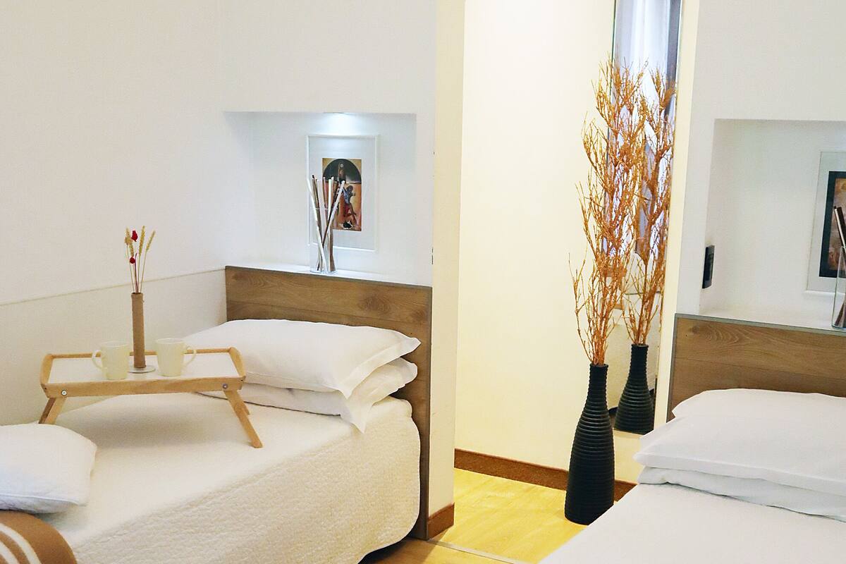 A hotel room with two beds, a wooden floor, white bedding, and two black vases with branches.