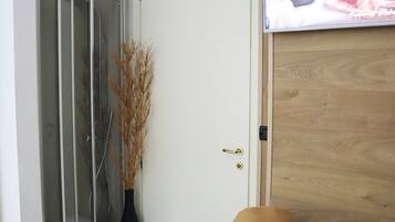 Een kamer met een douche, een zwarte stoel met kussen, een houten deur en een aan de muur bevestigde televisie.