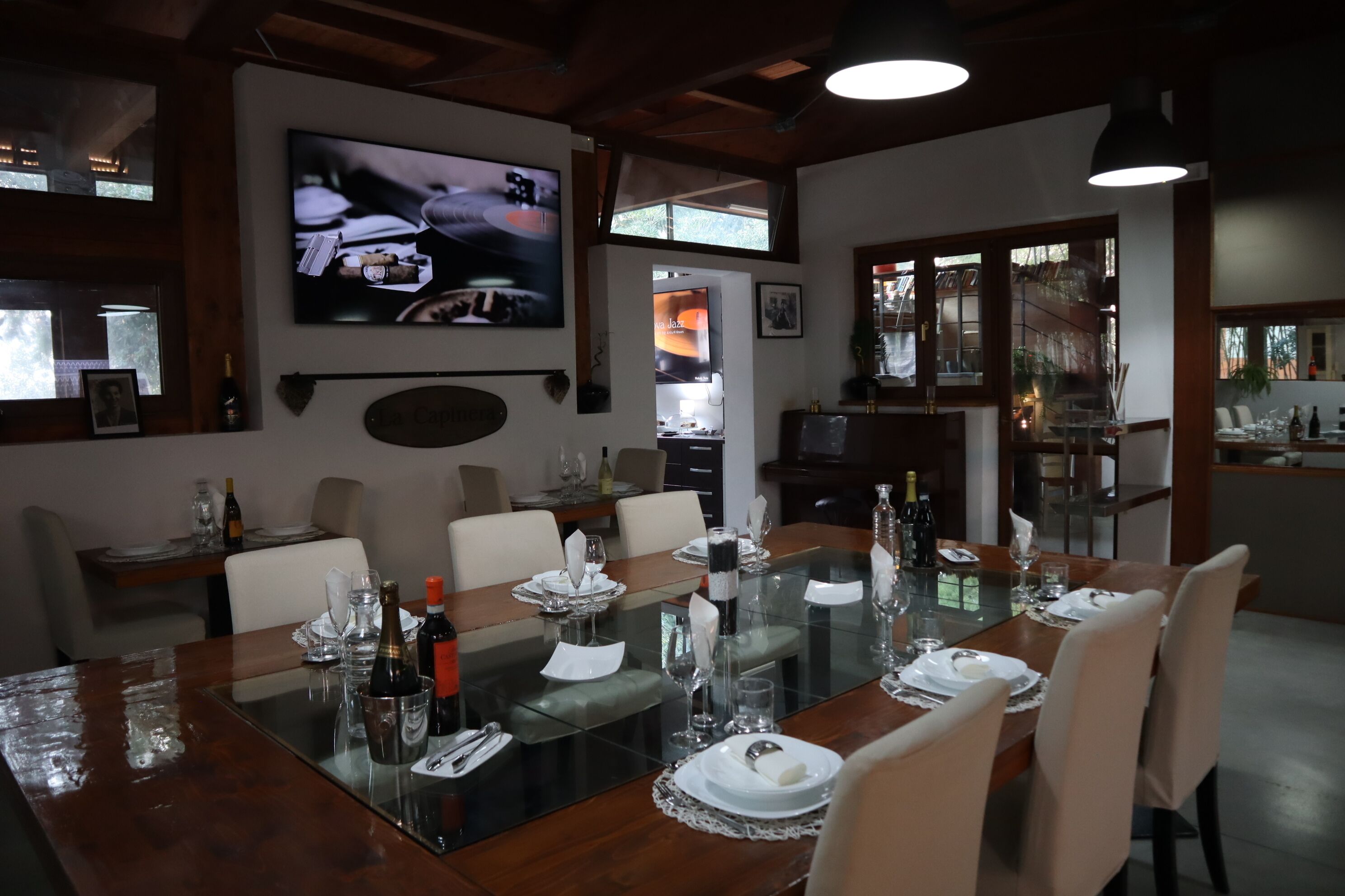 A dining area with a glass table set for six, surrounded by chairs. A television is mounted on the wall above the table.