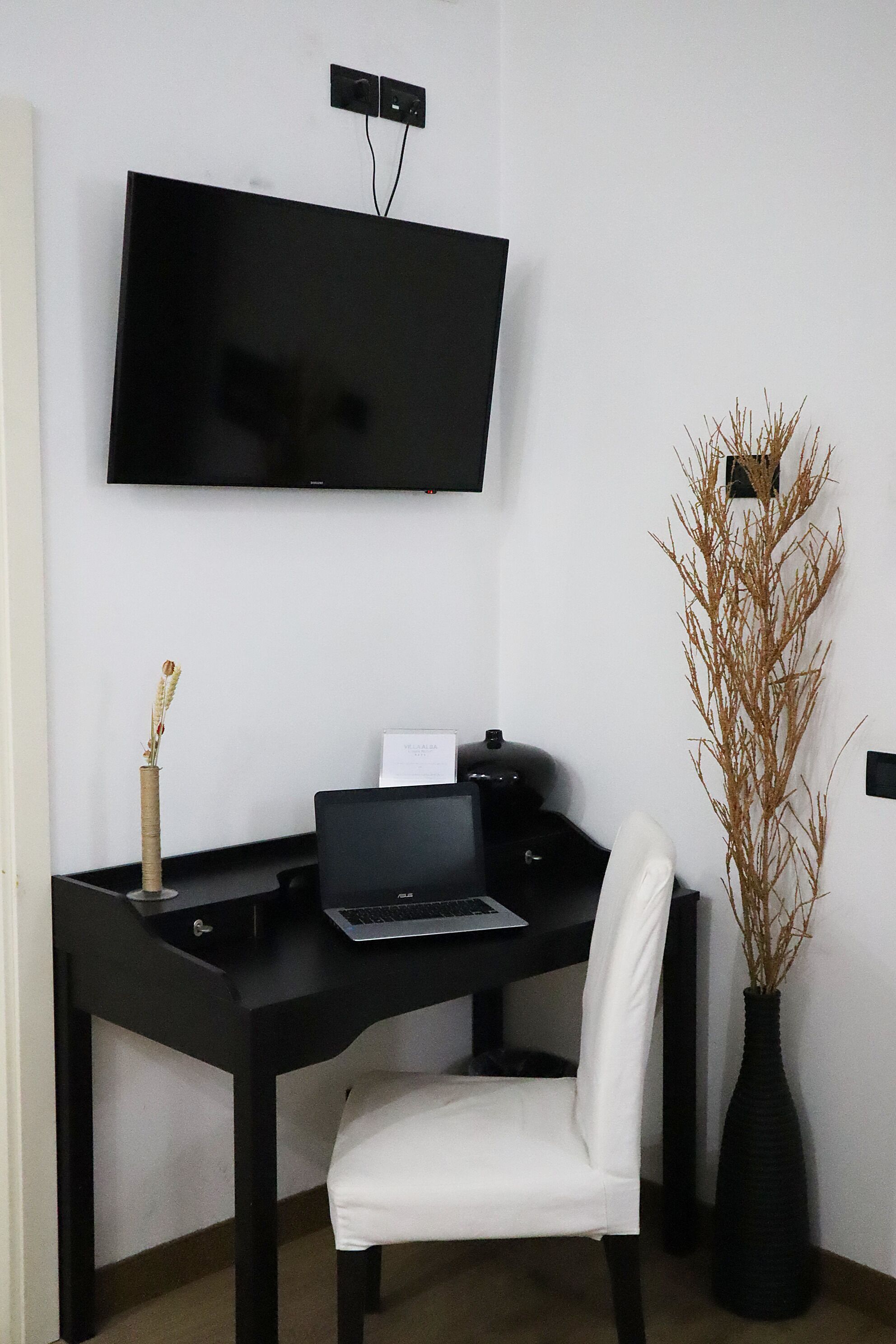 Un espai d'oficina modern amb escriptori negre, cadira blanca, un portàtil, televisió de pantalla plana penjada a la paret i un ramader decoratiu amb plantes seques.