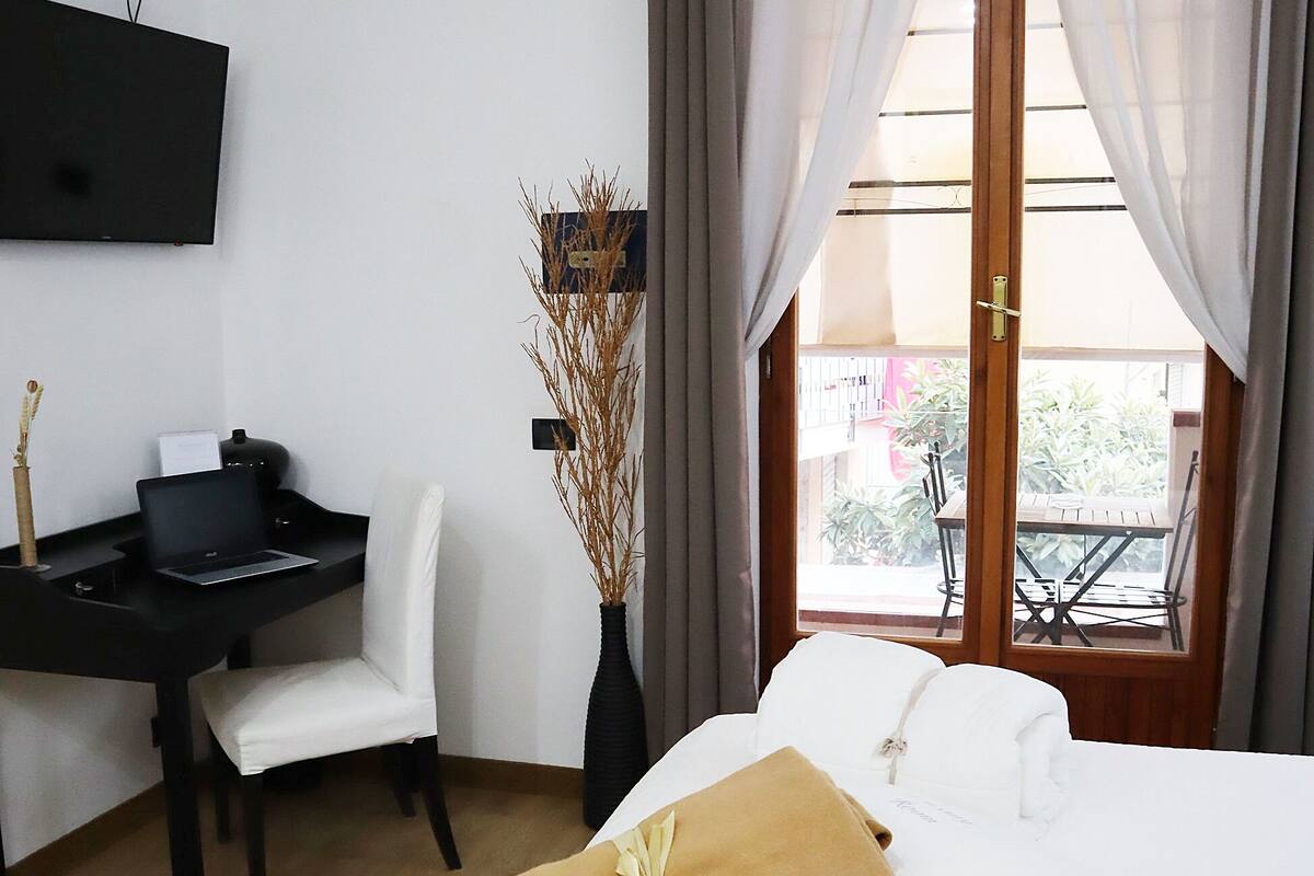 A hotel room with a bed, a desk with a laptop, a vase with branches, and a balcony with a table and chairs.