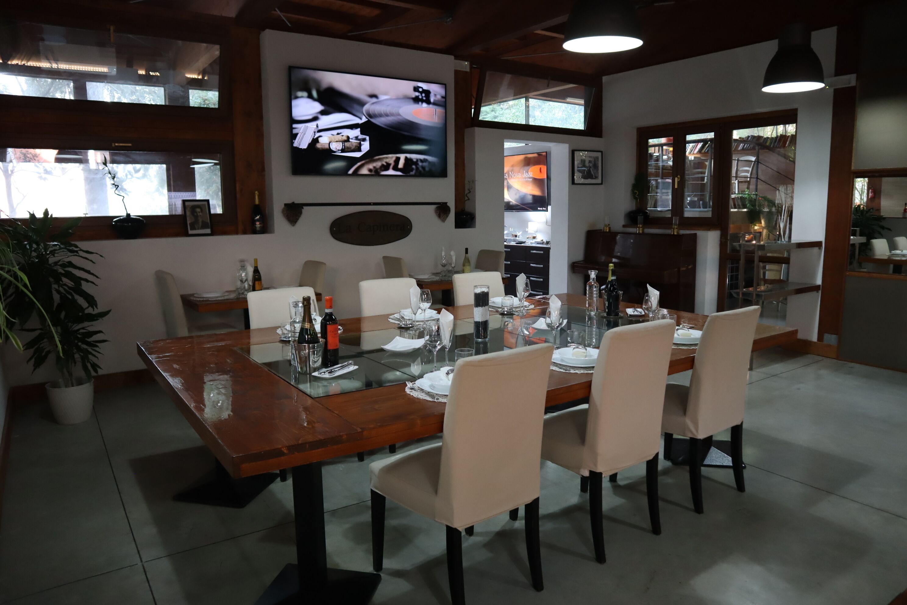 A dining area with a wooden table set for six, surrounded by chairs. A television is mounted on the wall above the table.