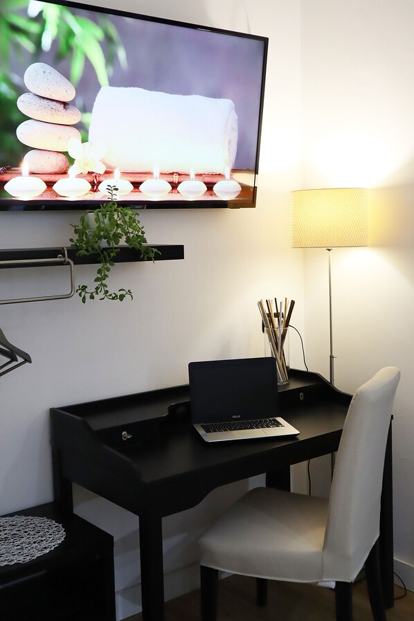 A modern workspace with a laptop on a black desk, a white chair, a TV displaying a serene spa scene, a floor lamp, and a small plant.