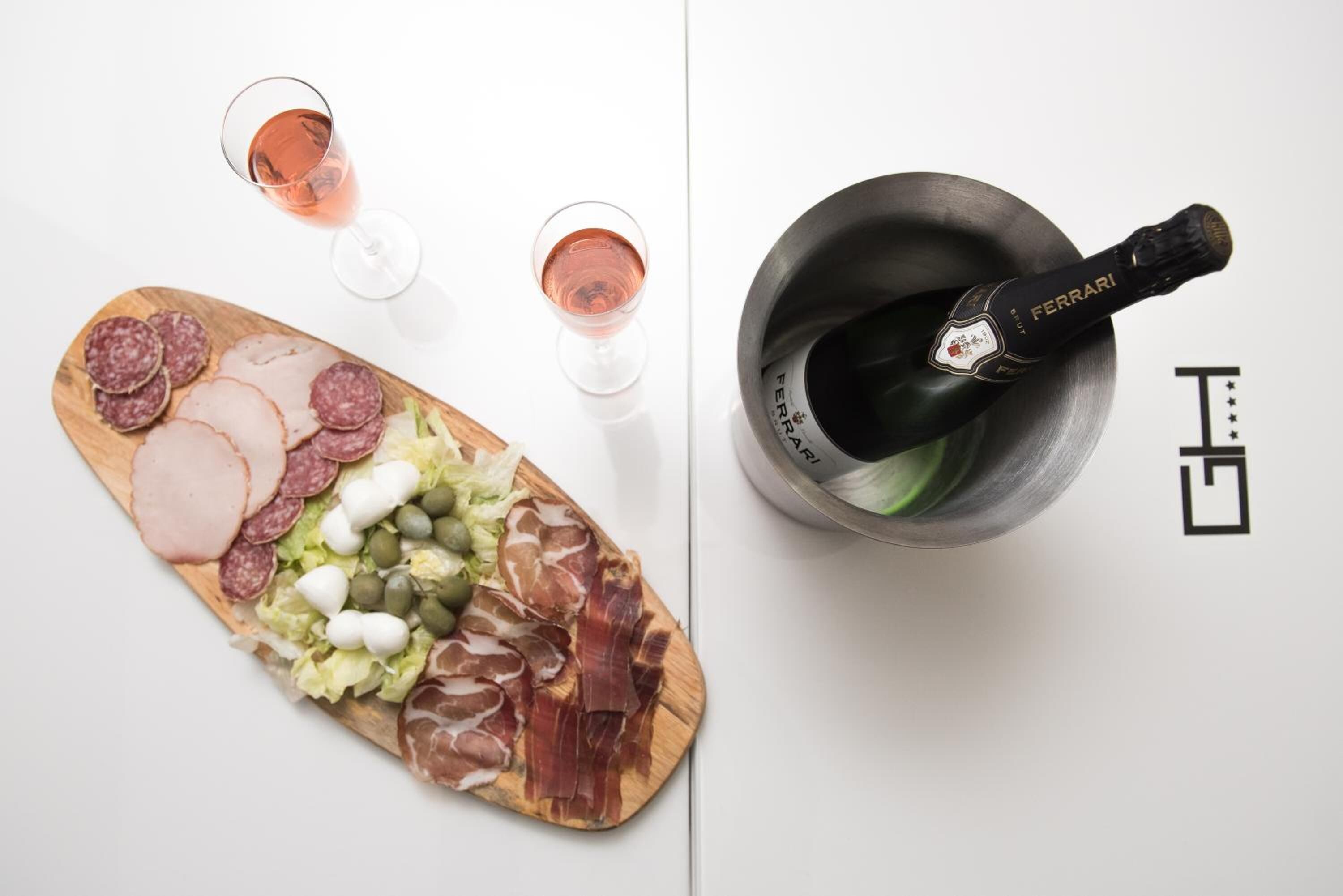 Une planche de charcuterie avec divers types de viandes et de fromages, deux verres de vin rosé et une bouteille de champagne Ferrari dans une carafe.