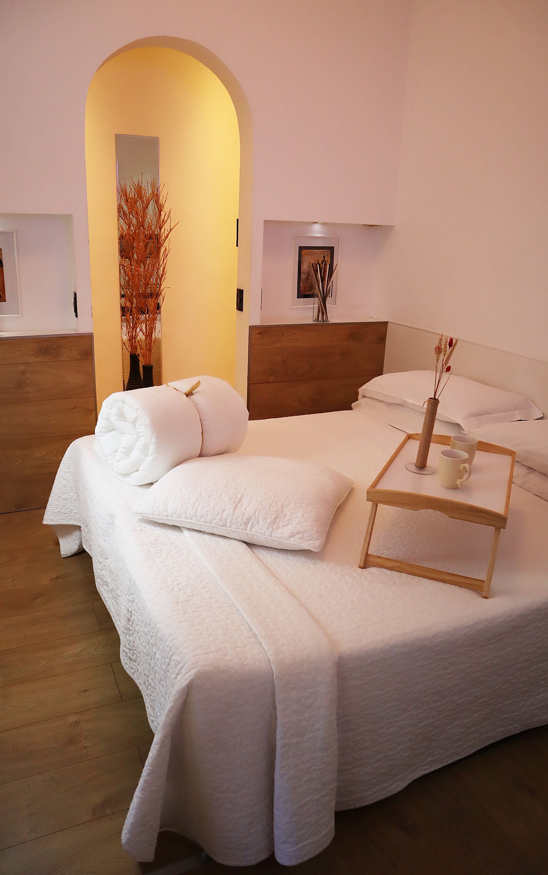 A neatly made bed with white linens, a wooden headboard, and a bedside table with a vase and two cups.