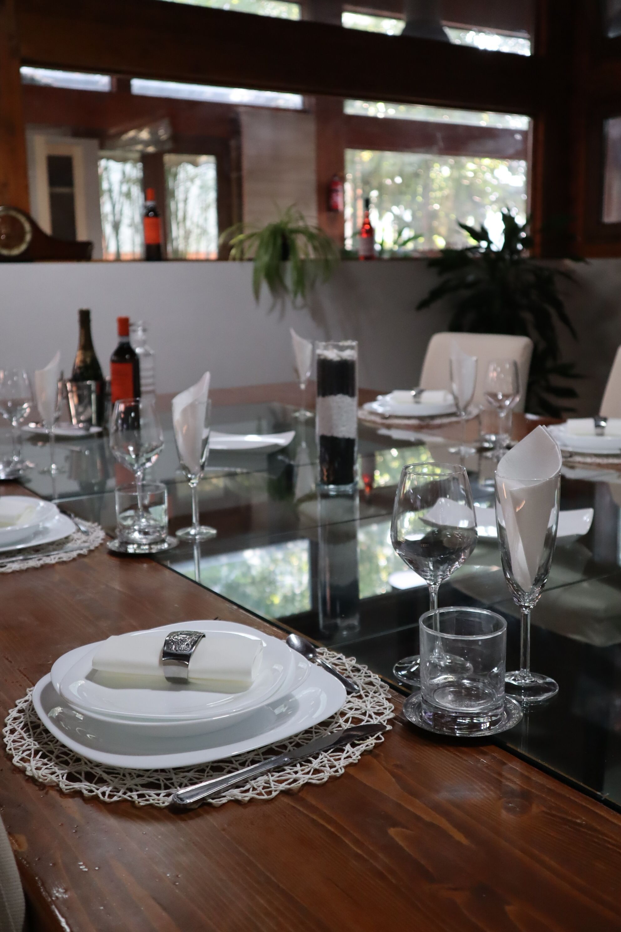 Une table à manger dressée avec des assiettes, des verres et une bouteille de vin, donnant sur l’extérieur par une fenêtre.