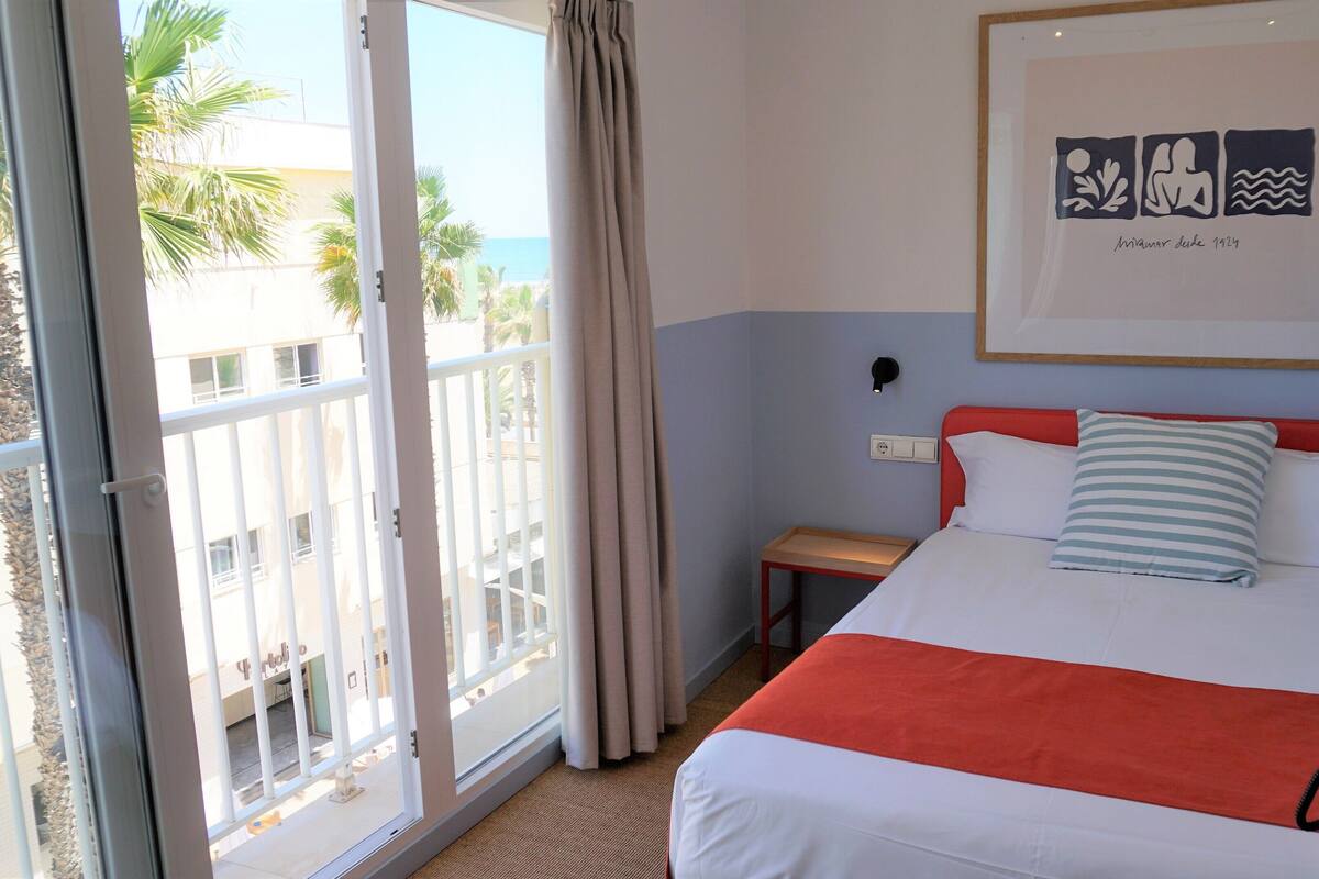 Hotelkamer met een bed, nachtkastje, een ingelijst beeld en uitzicht op palmbomen en een gebouw.