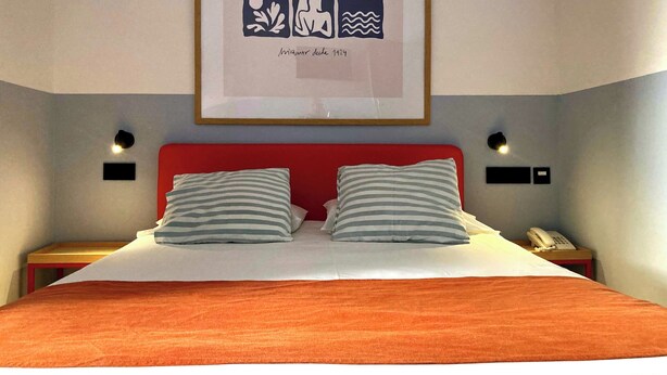 Une chambre d’hôtel avec une tête de lit rouge, deux coussins à rayures, une œuvre d’art encadrée au-dessus du lit et des lampes de chevet.