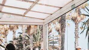 A restaurant with a large glass-enclosed dining area, wooden furniture, and a view of palm trees. - Hotel Miramar Valencia (Valencia)