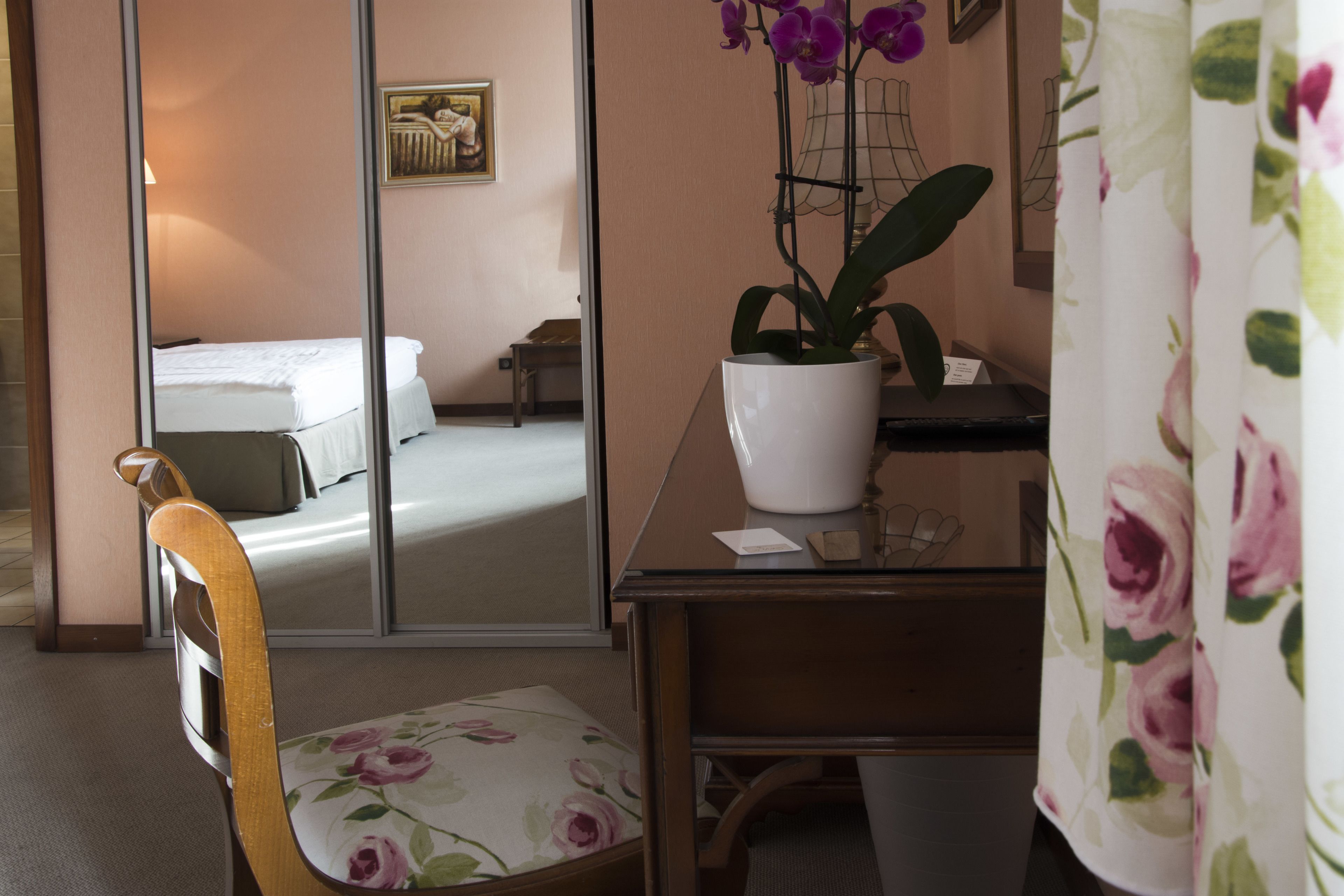 Habitación de hotel con cama, un escritorio con una orquídea en maceta, una silla con cojín floral y un espejo que refleja la habitación.