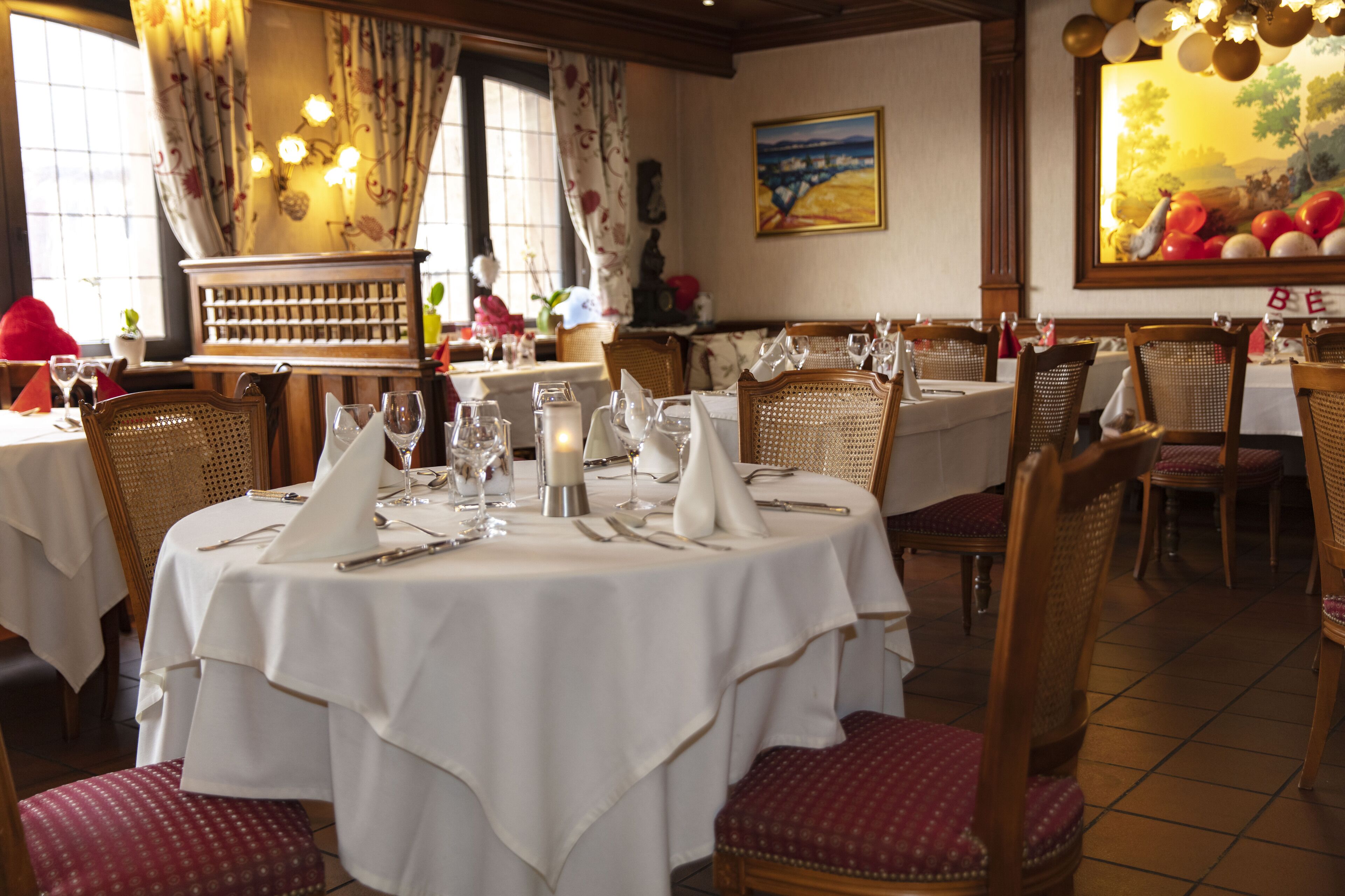 Een restaurant met ronde tafels gedekt voor diners, rieten stoelen en een groot schilderij aan de muur.