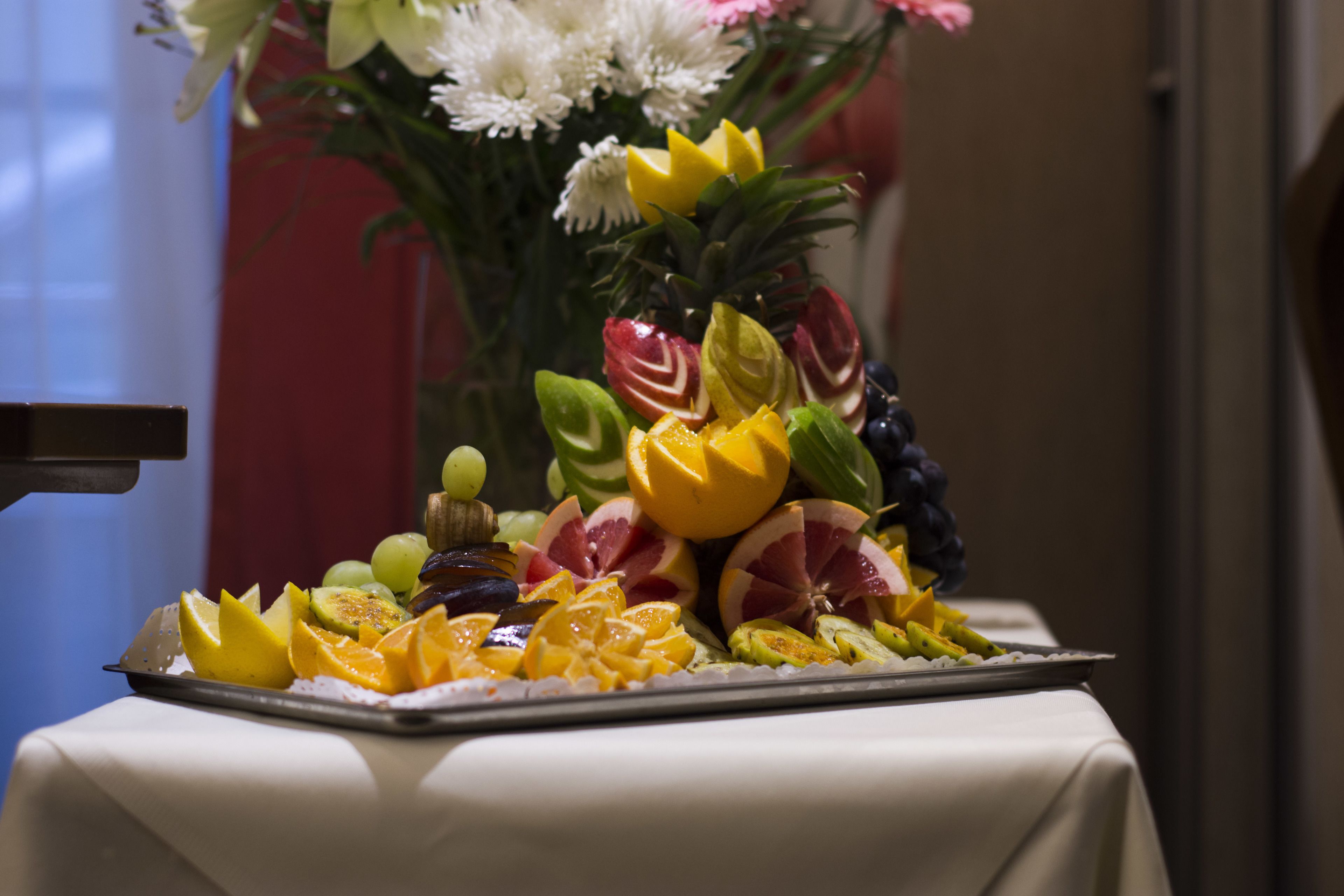 A fruit platter on a table with a white tablecloth, surrounded by a vase of flowers.