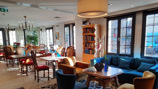 A well-lit hotel lobby with a dining area, seating arrangements, and a bookshelf. - Hostellerie Saint Florent (Oberhaslach)