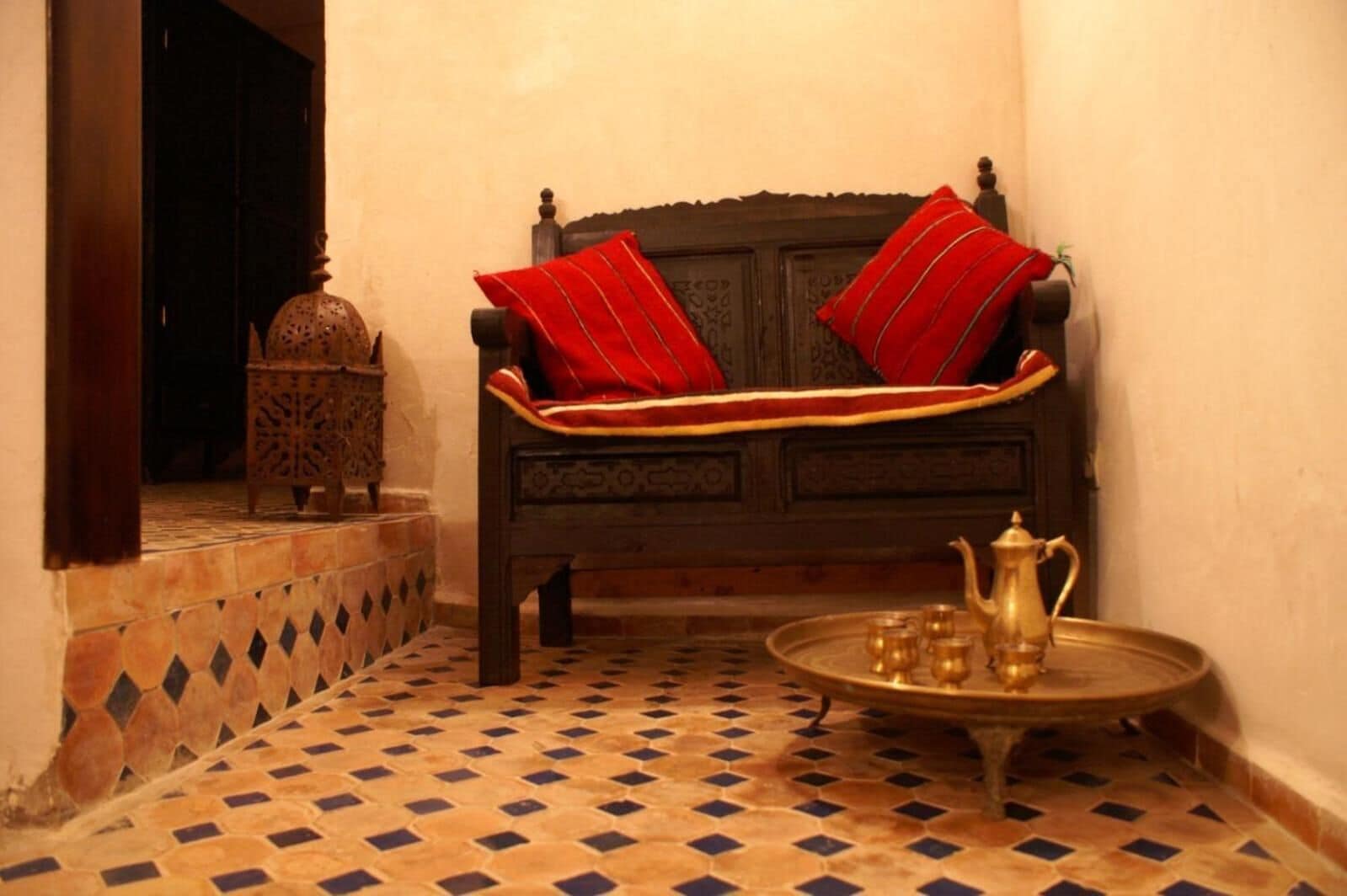 Una habitación tradicional marroquí con un sofá de madera, cojines rojos, suelo estampado y una bandeja de latón con tetera y tazas.
