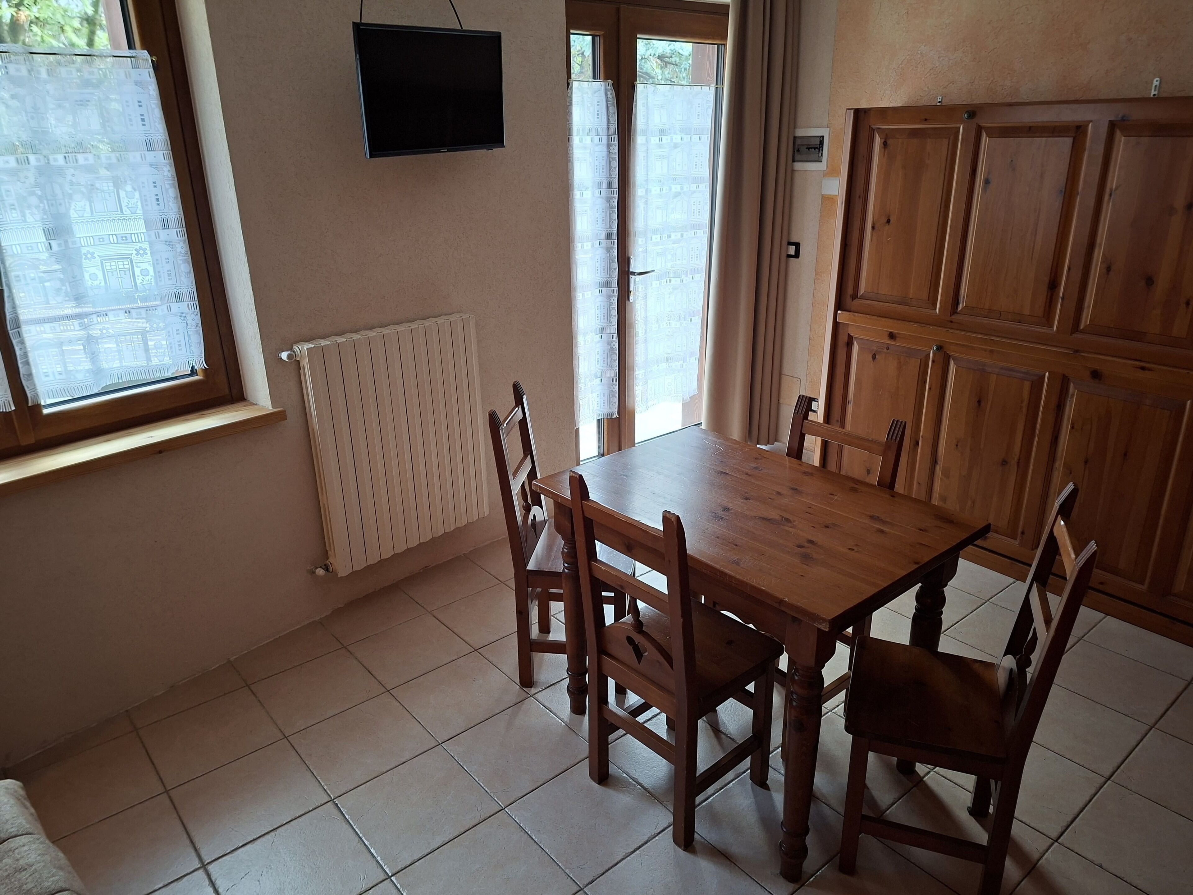 A dining area with a wooden table and chairs, a TV mounted on the wall, and a window with a view.