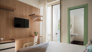 A hotel room with a bed, a TV mounted on the wall, a small table with a chair, and a view of the outside balcony. - Hotel Calypso (Rimini)