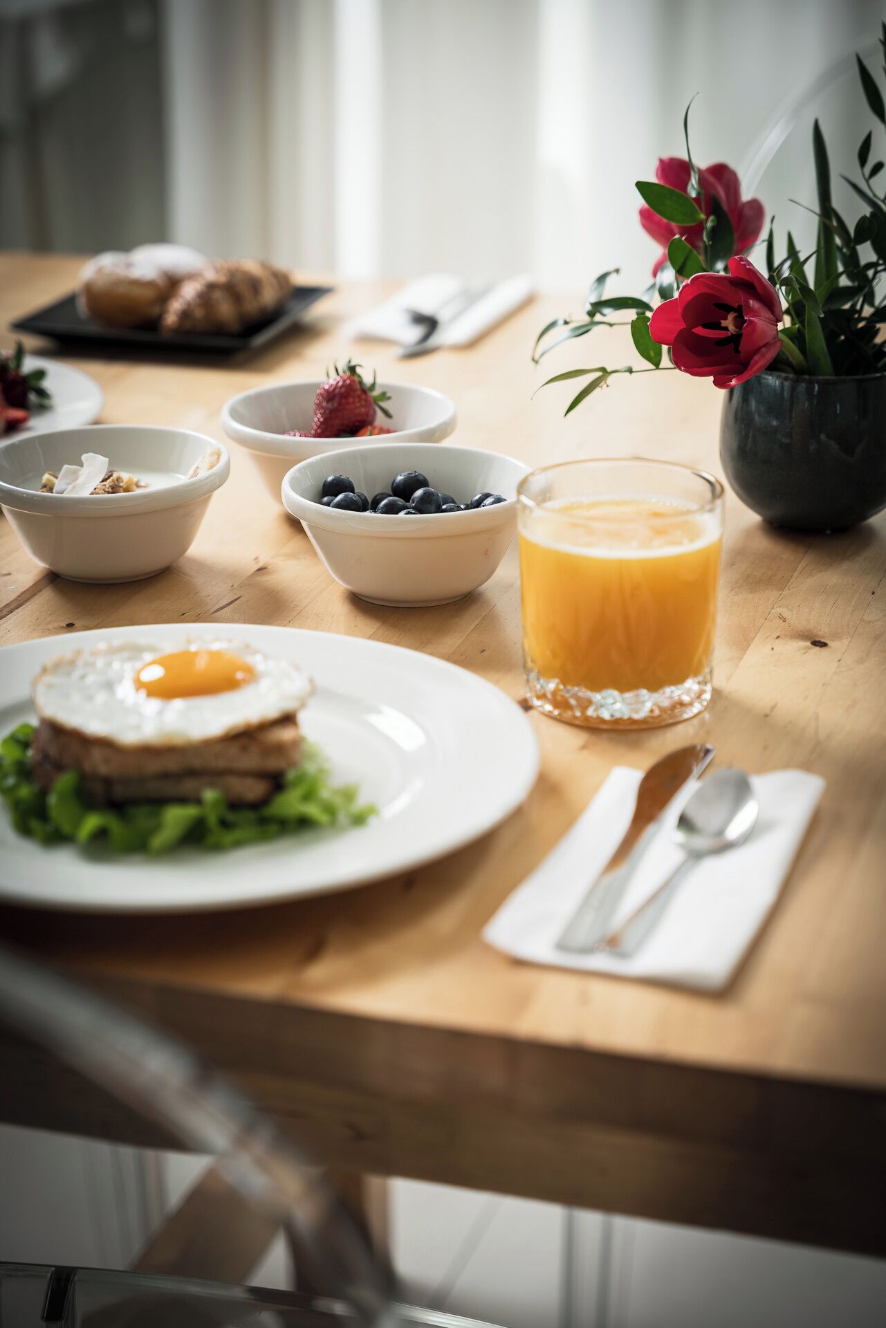A breakfast setting with a plate of eggs, a glass of orange juice, bowls of berries, and a vase of flowers on a wooden table.