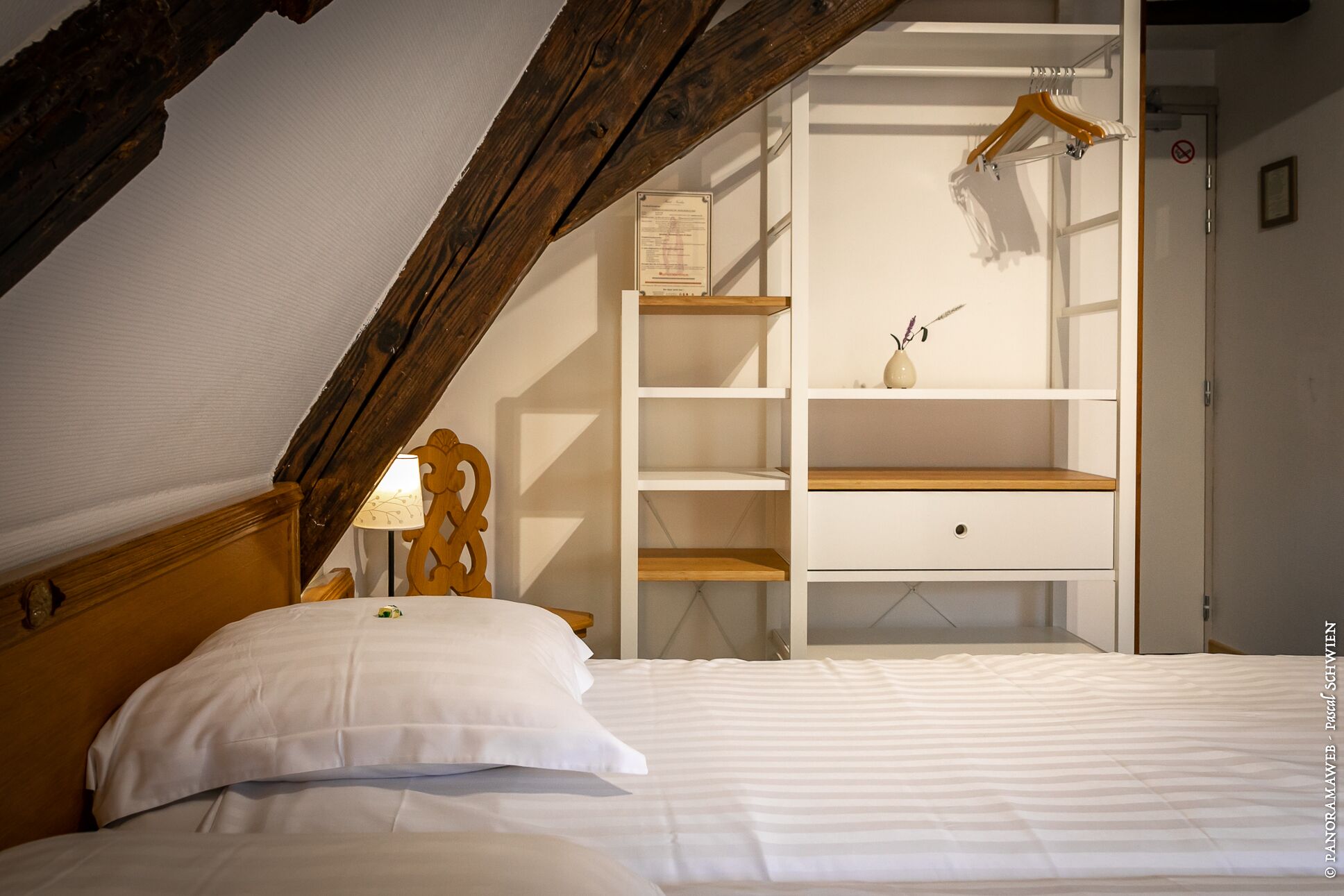 A bedroom with a sloped ceiling, a bed with white bedding, a wooden headboard, a bedside table with a lamp, and a white wardrobe with shelves and a hanging rod.