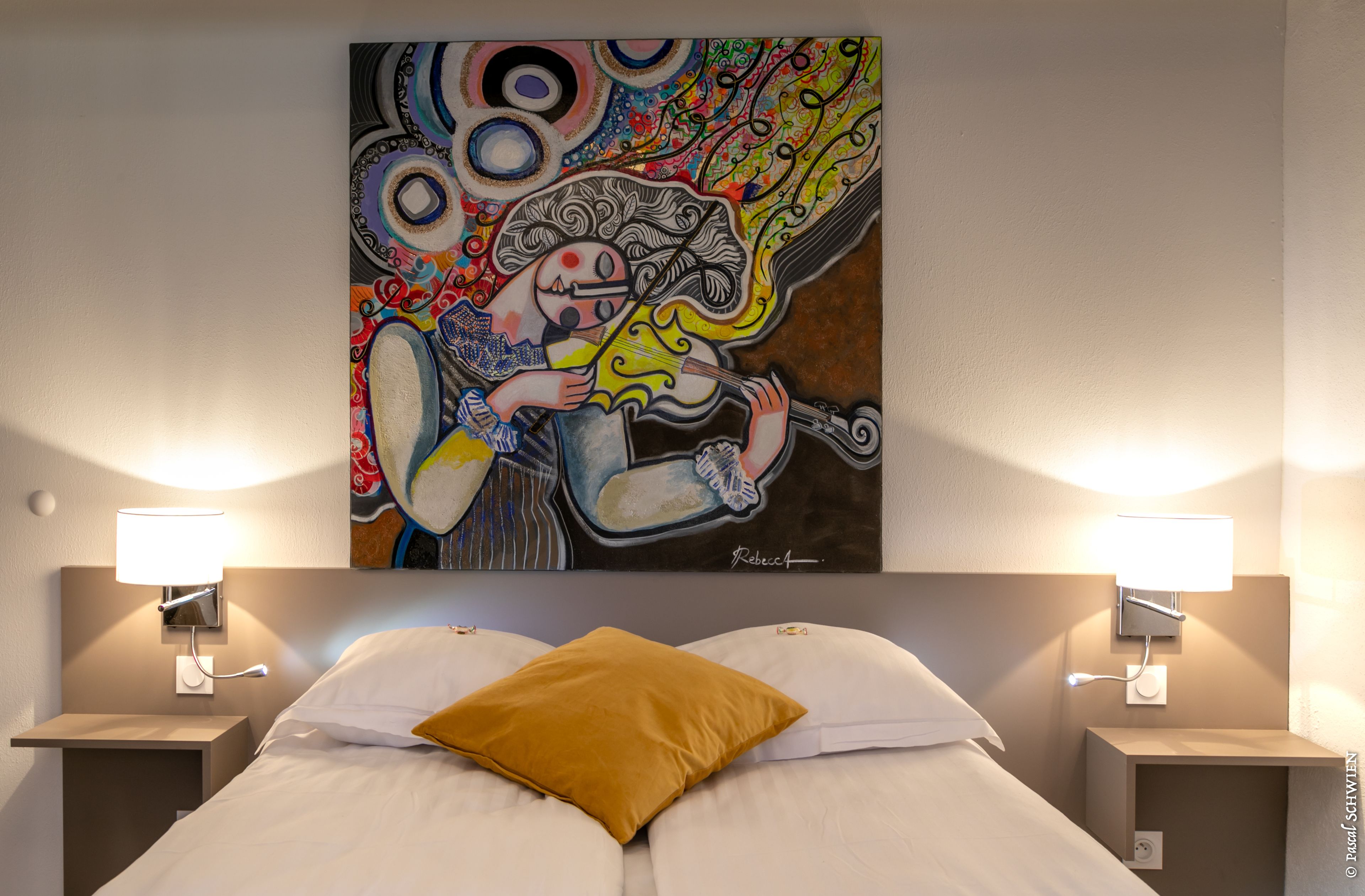 Ein Hotelzimmer mit Bett, Nachttisch, Lampe und einem farbenfrohen, abstrakten Gemälde an der Wand.