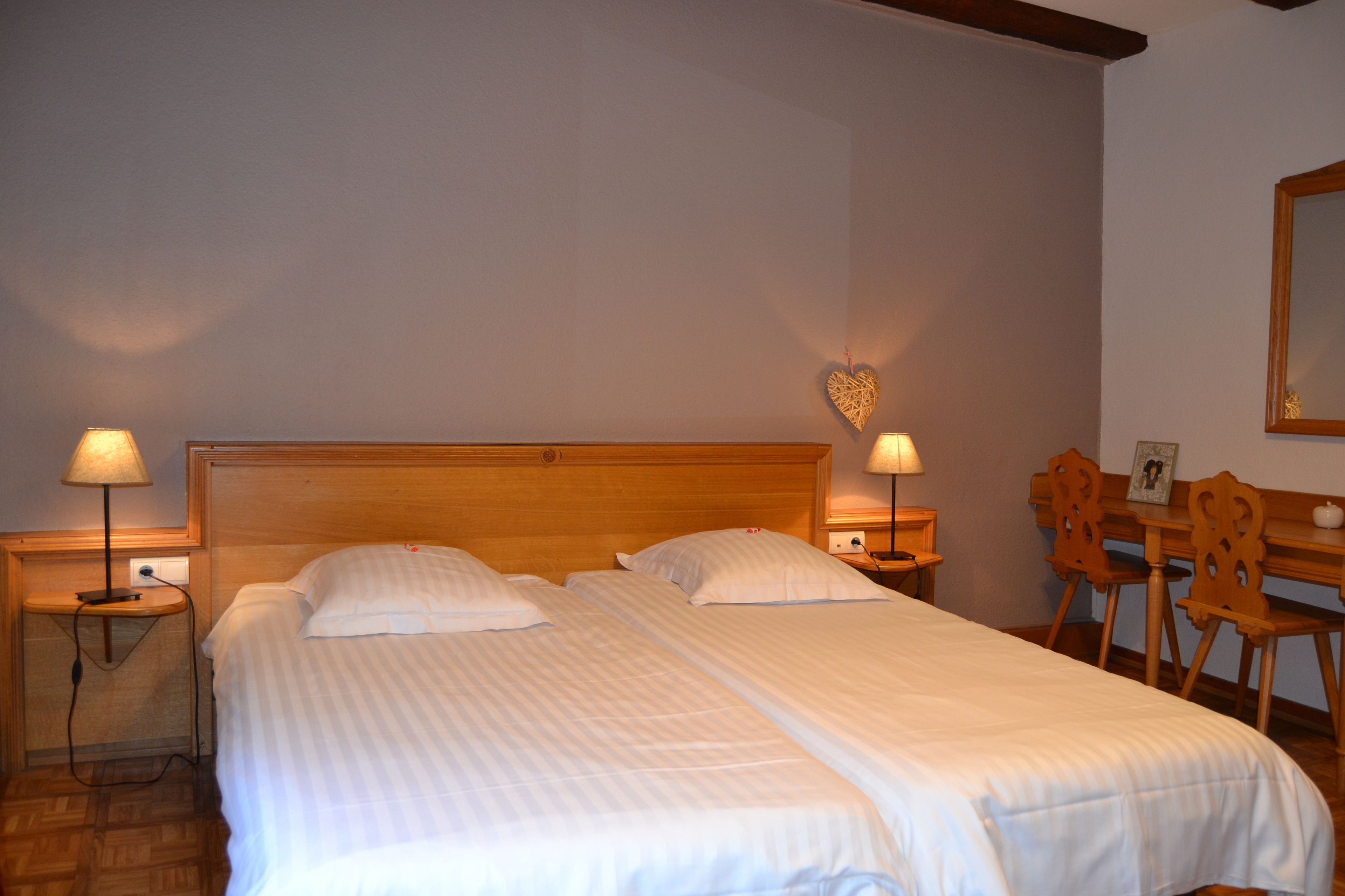 Uma cama de casal com duas mesinhas de cabeceira, uma cabeceira de madeira, uma decoração de coração na parede e uma escrivaninha de madeira com cadeiras.