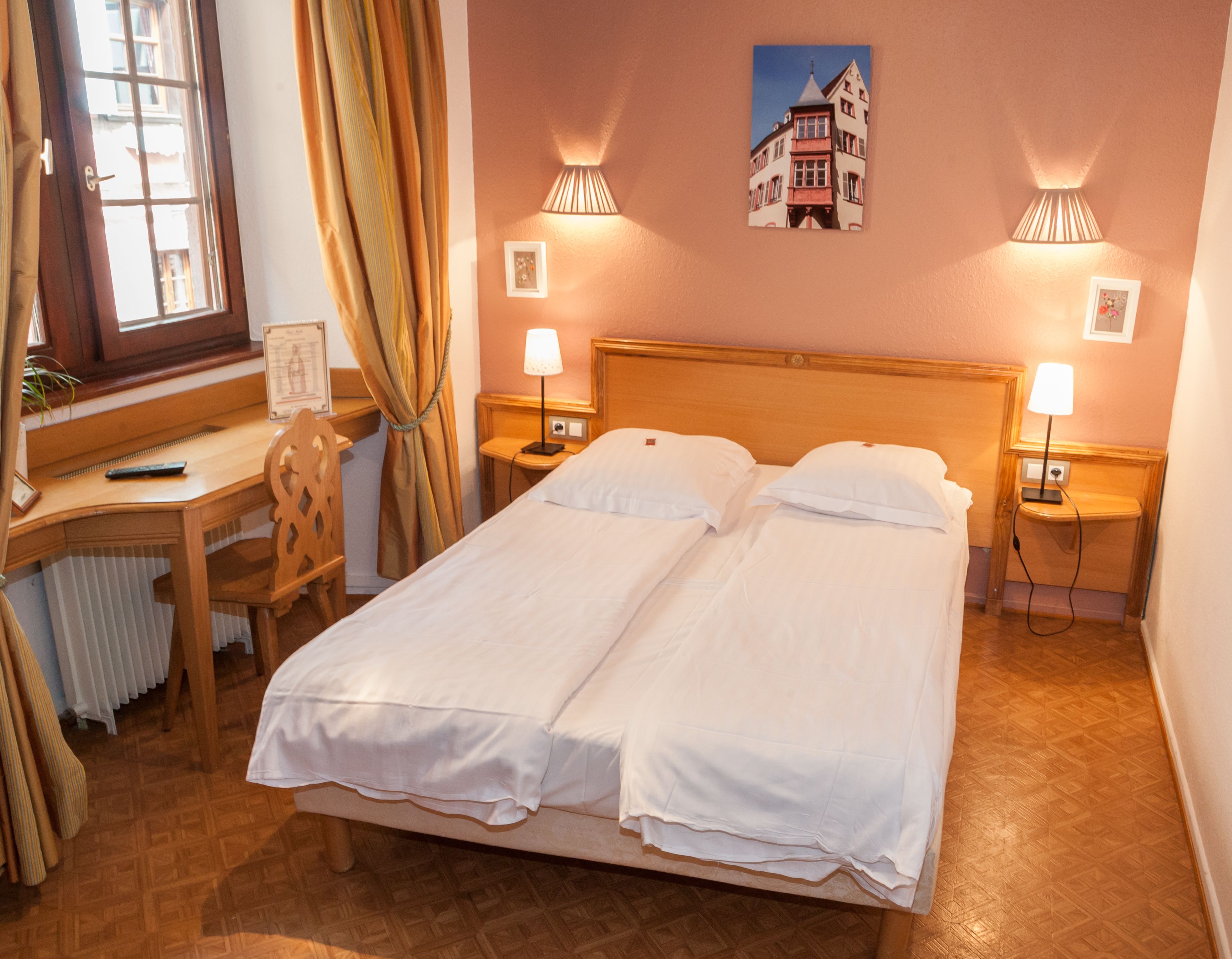Un lit double avec du linge de lit blanc, un bureau en bois avec une chaise, deux lampes de chevet et un tableau représentant un bâtiment accroché au mur.