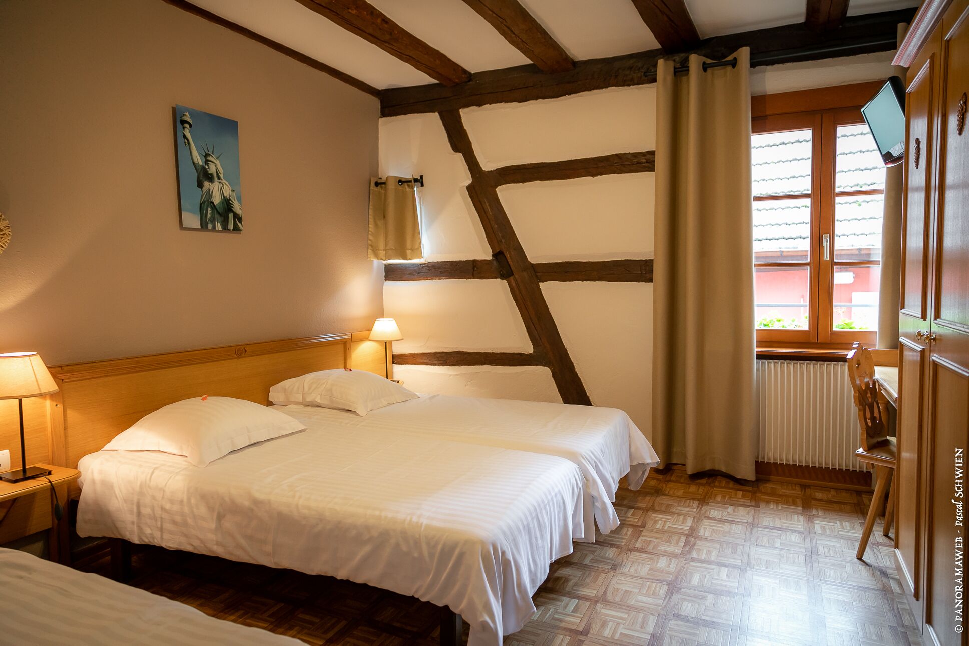 Een hotelkamer met twee bedden, een houten plafond, een schilderij van de Vrijheidsbeeld en een raam met gordijnen, plus een bureau met stoel.