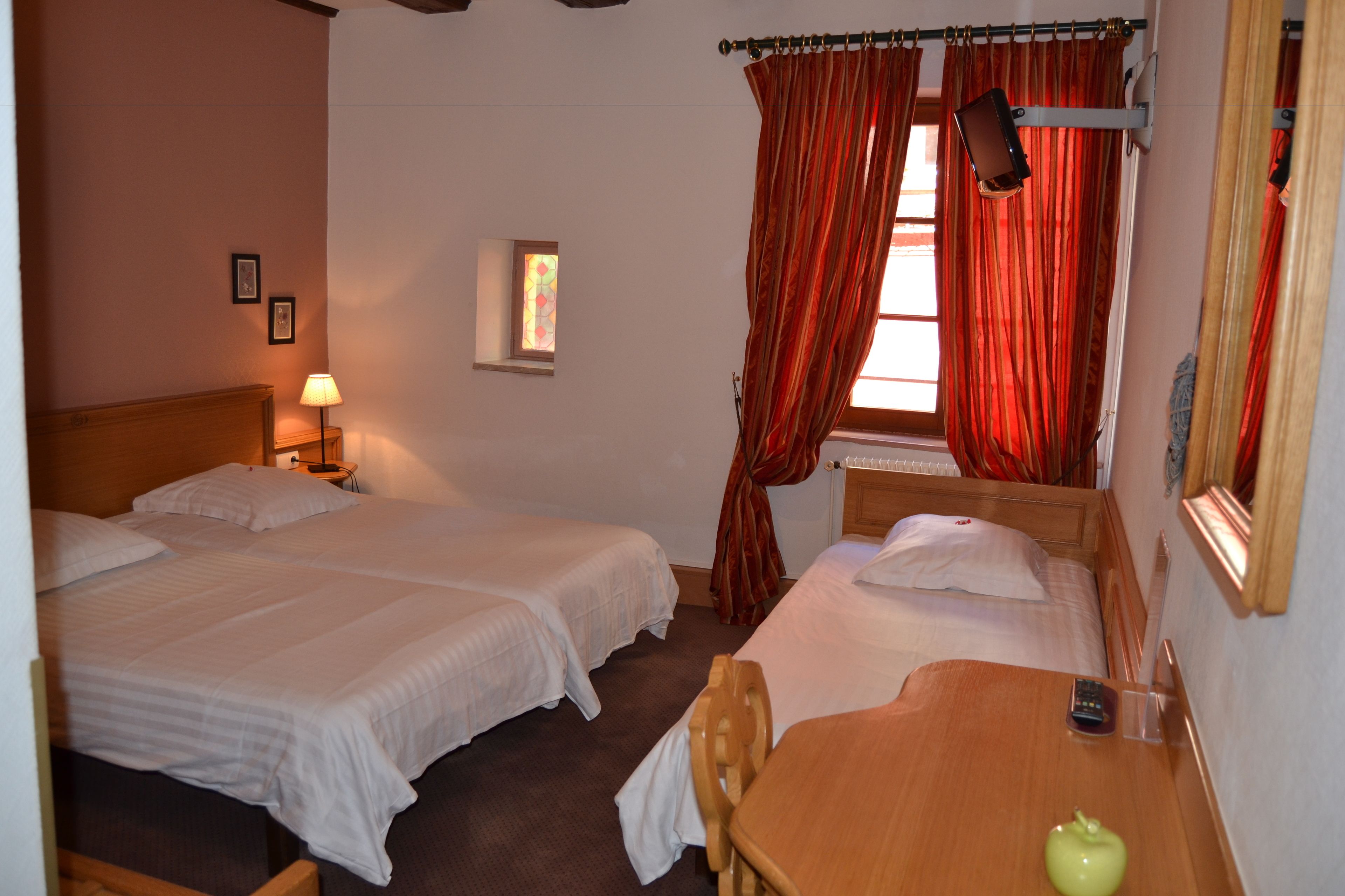 A hotel room with two single beds, a wooden table, a chair, a small window with curtains, and a wall-mounted lamp.