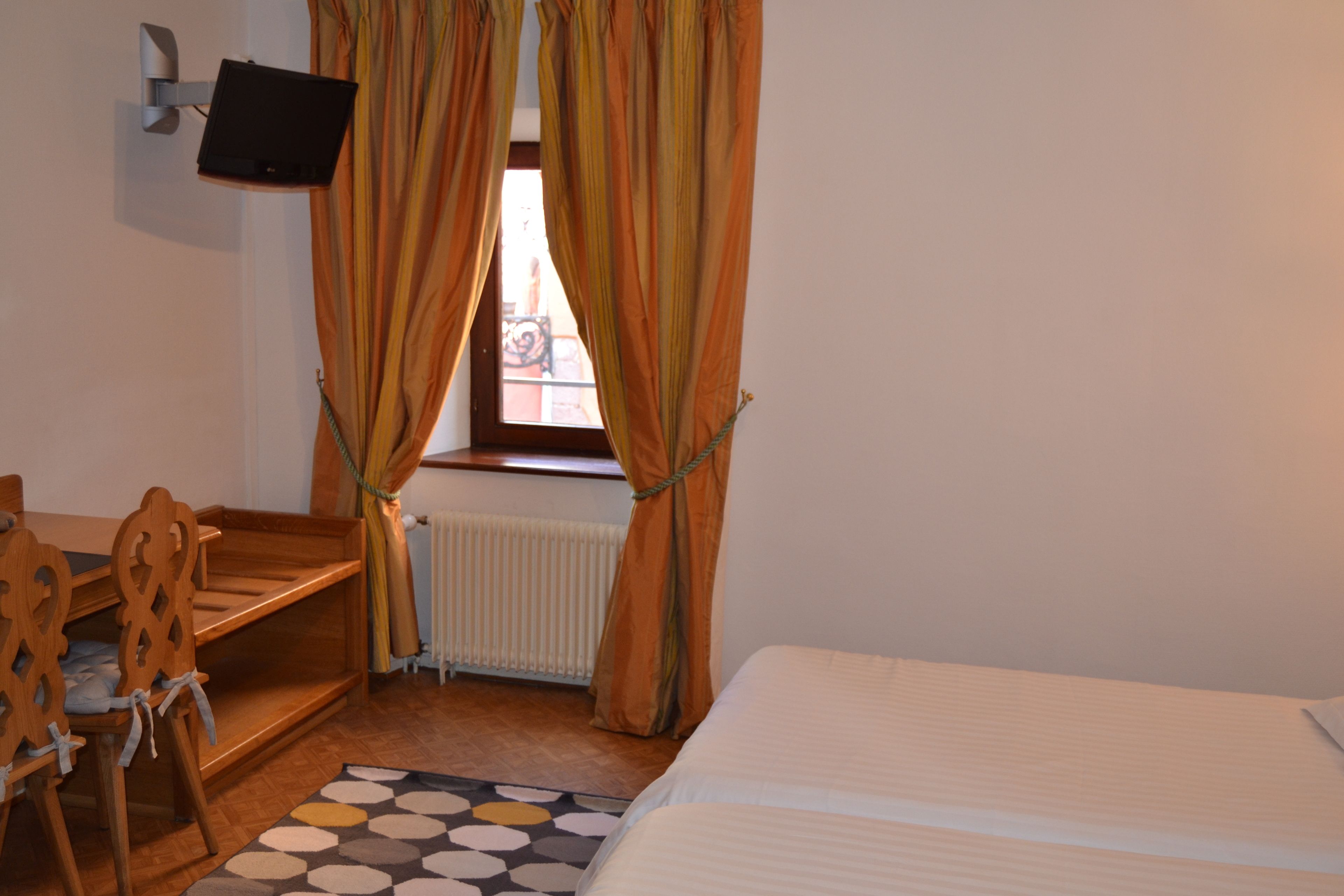 A room with a bed, a desk, and a chair. There is a window with curtains and a television mounted on the wall.