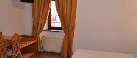 A room with a bed, a desk, and a chair. There is a window with curtains and a television mounted on the wall.