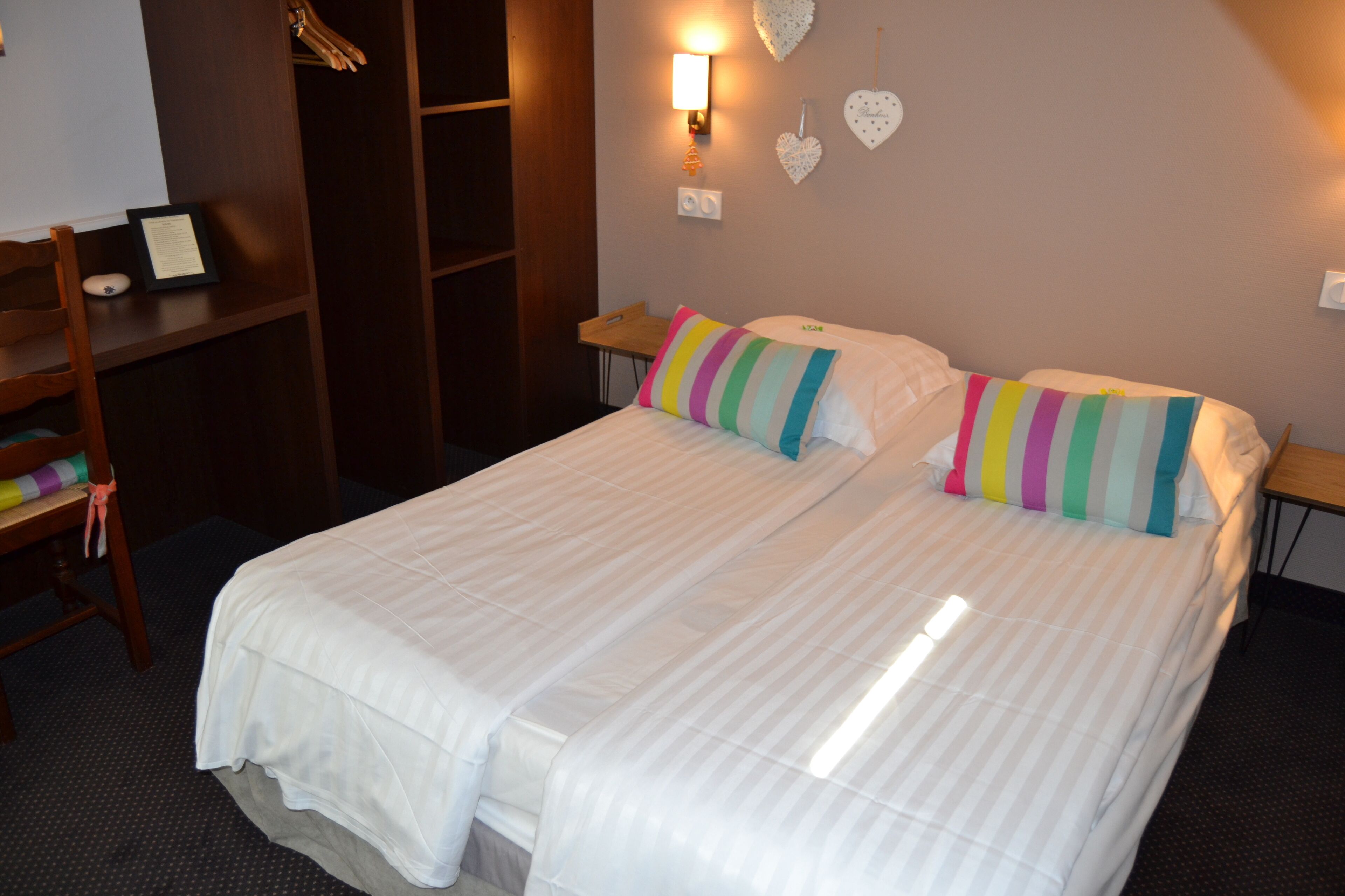 A double bed with striped bedding and colorful pillows, a wooden chair, a small table, and a wardrobe.