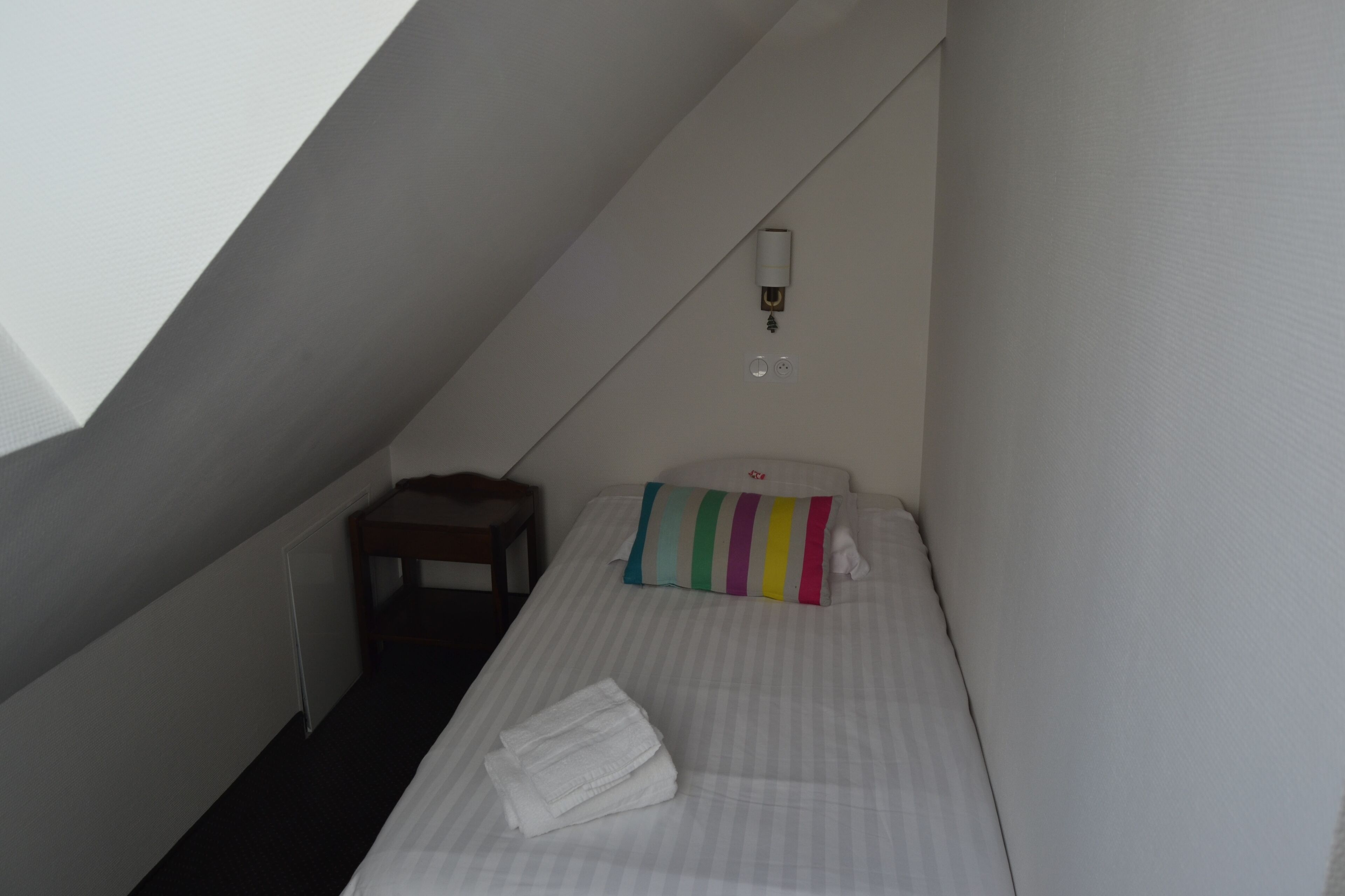 Una habitación pequeña y estrecha con una cama individual, una mesita y una lámpara fijada a la pared.