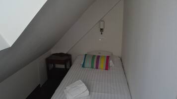 Una habitación pequeña y estrecha con una cama individual, una mesita y una lámpara fijada a la pared.