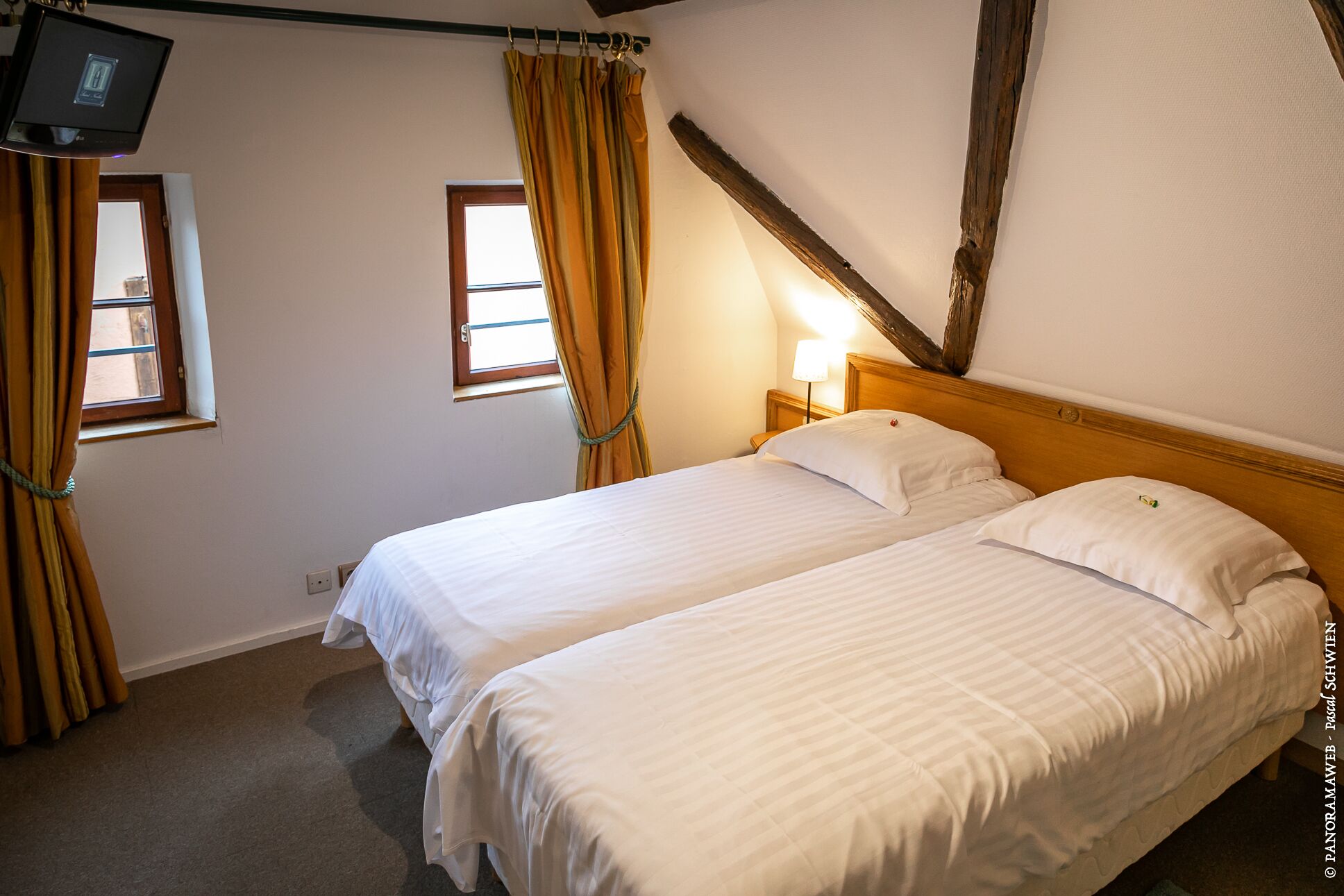 Una habitación con dos camas individuales, un televisor fijado a la pared y dos ventanas con cortinas.