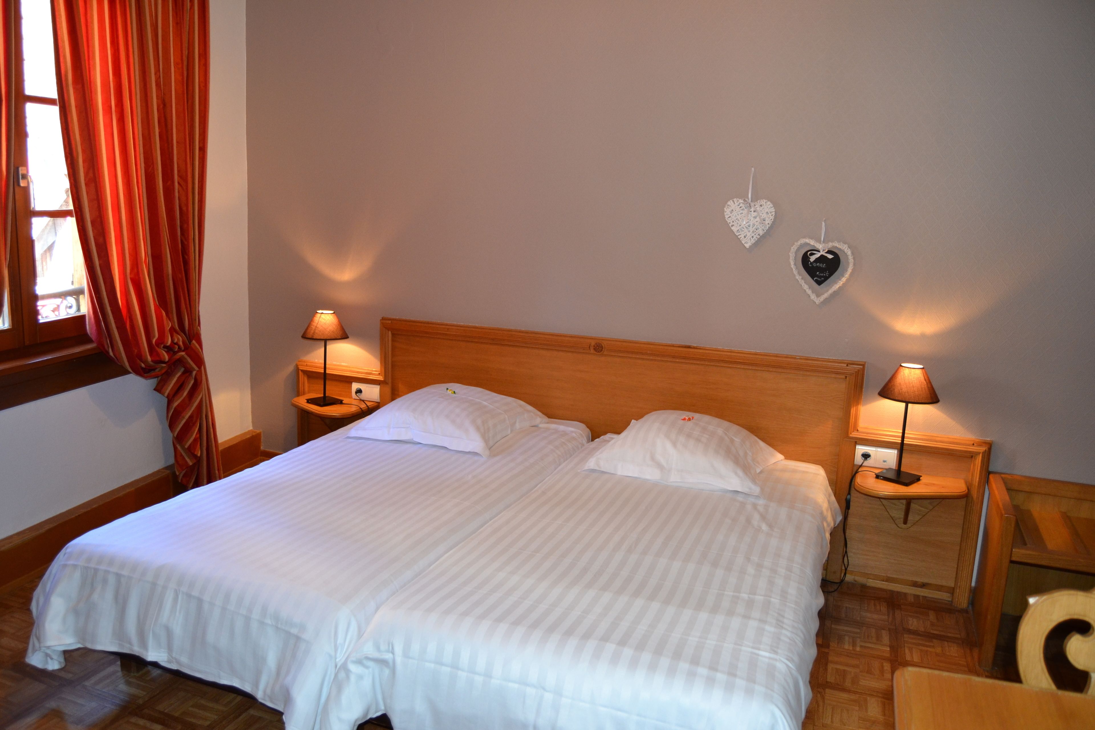 A double bed with two nightstands, each with a lamp, in a room with wooden flooring and a window with curtains.