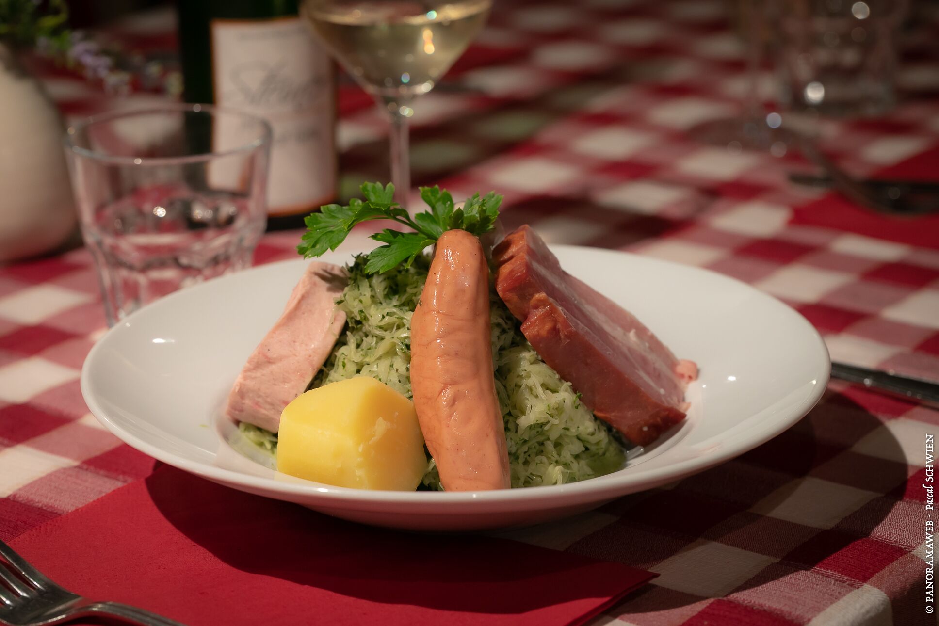 Une assiette contenant des saucisses, de la choucroute et une pomme de terre bouillie, le tout posé sur une nappe à carreaux.
