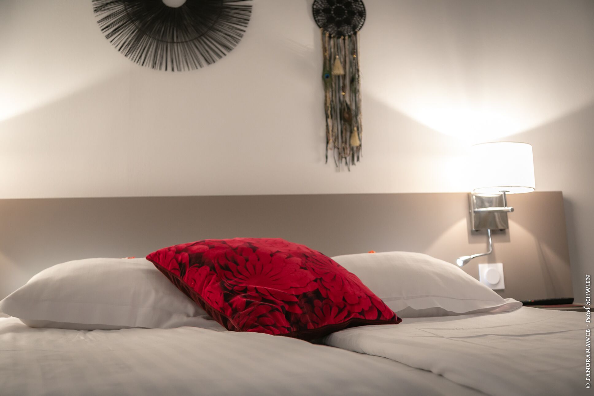 Een net opgemaakt bed met witte en rode kussens, een wandlamp en een dreamcatcher.