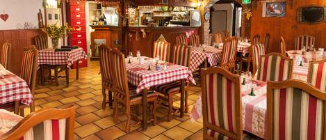 A restaurant with wooden tables and chairs, a kitchen in the background, and a tiled floor.