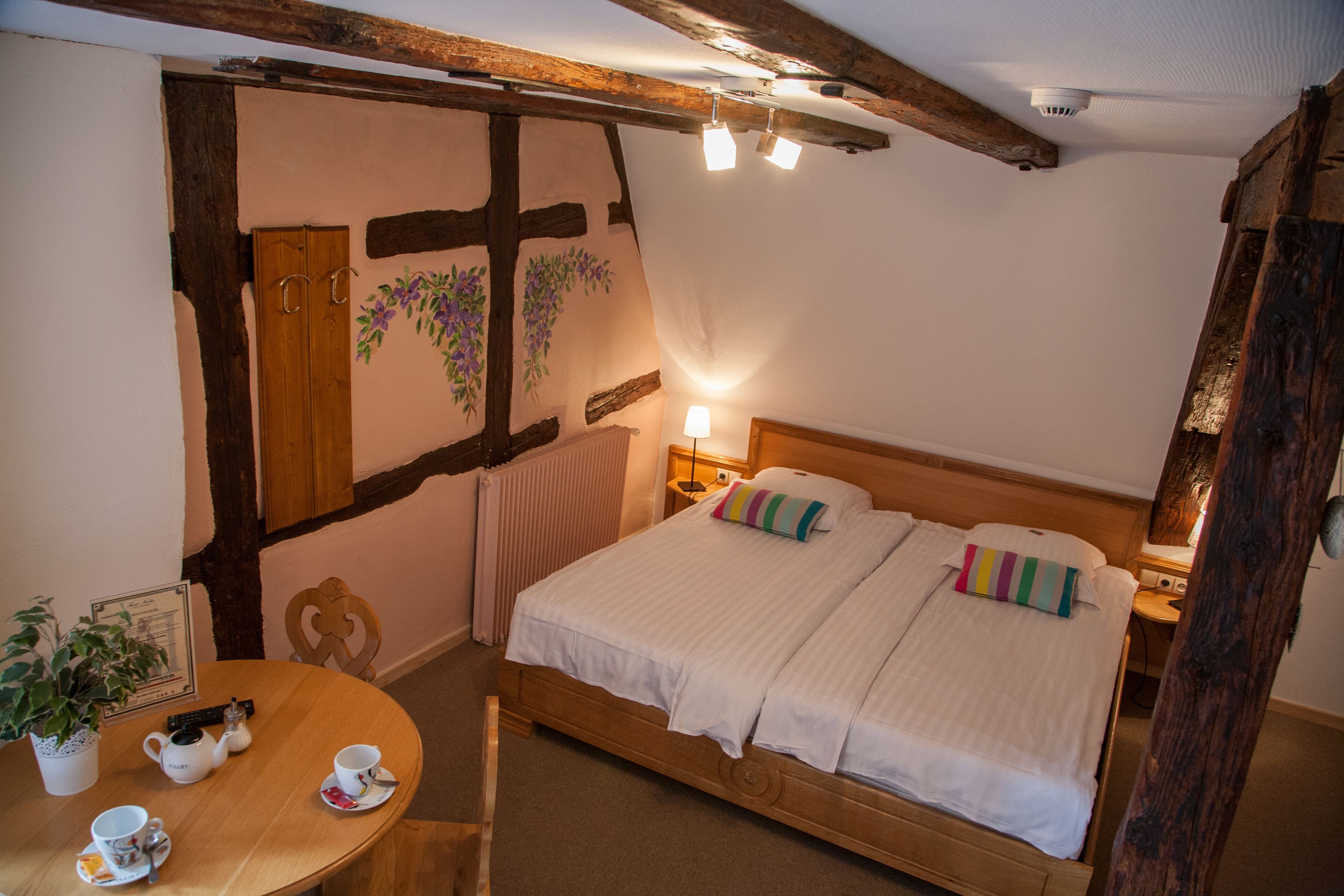 A bedroom with a wooden bed, a bedside table with a lamp, a chair, and a small table with a teapot and cups.