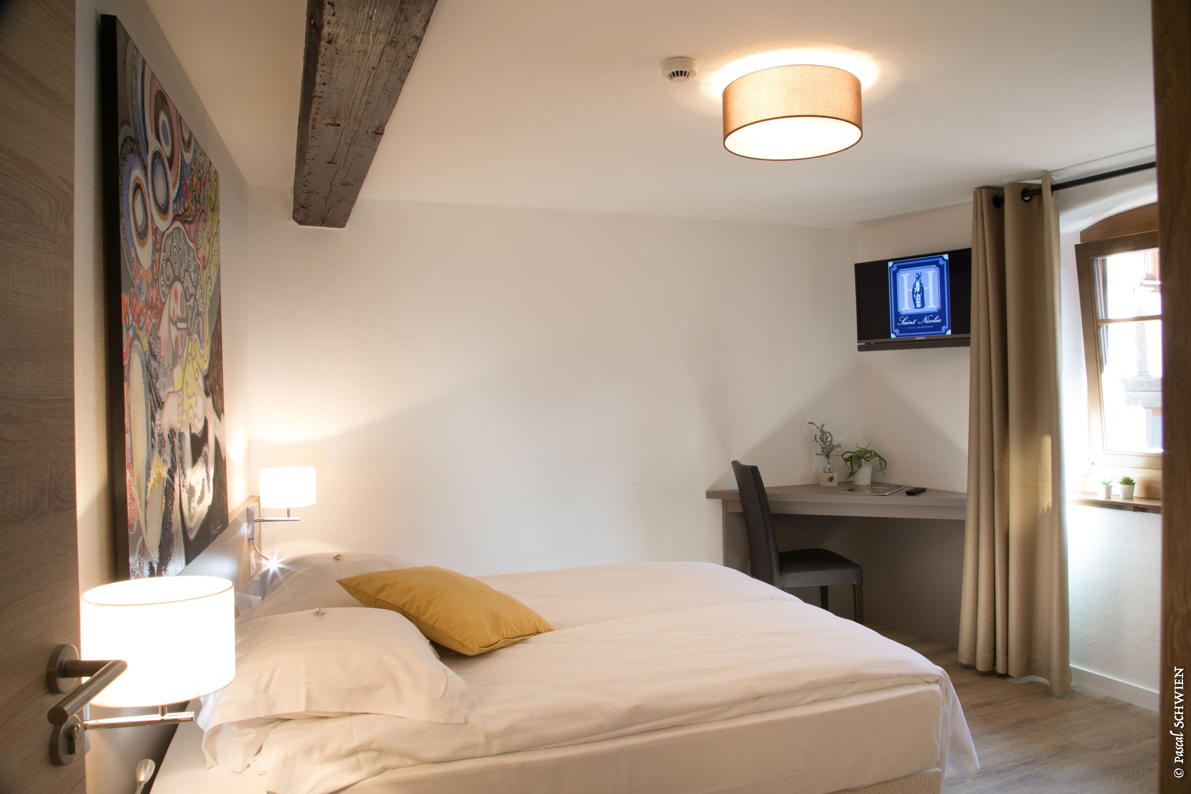 A hotel room with a bed, a desk, a chair, a TV, and a painting on the wall.