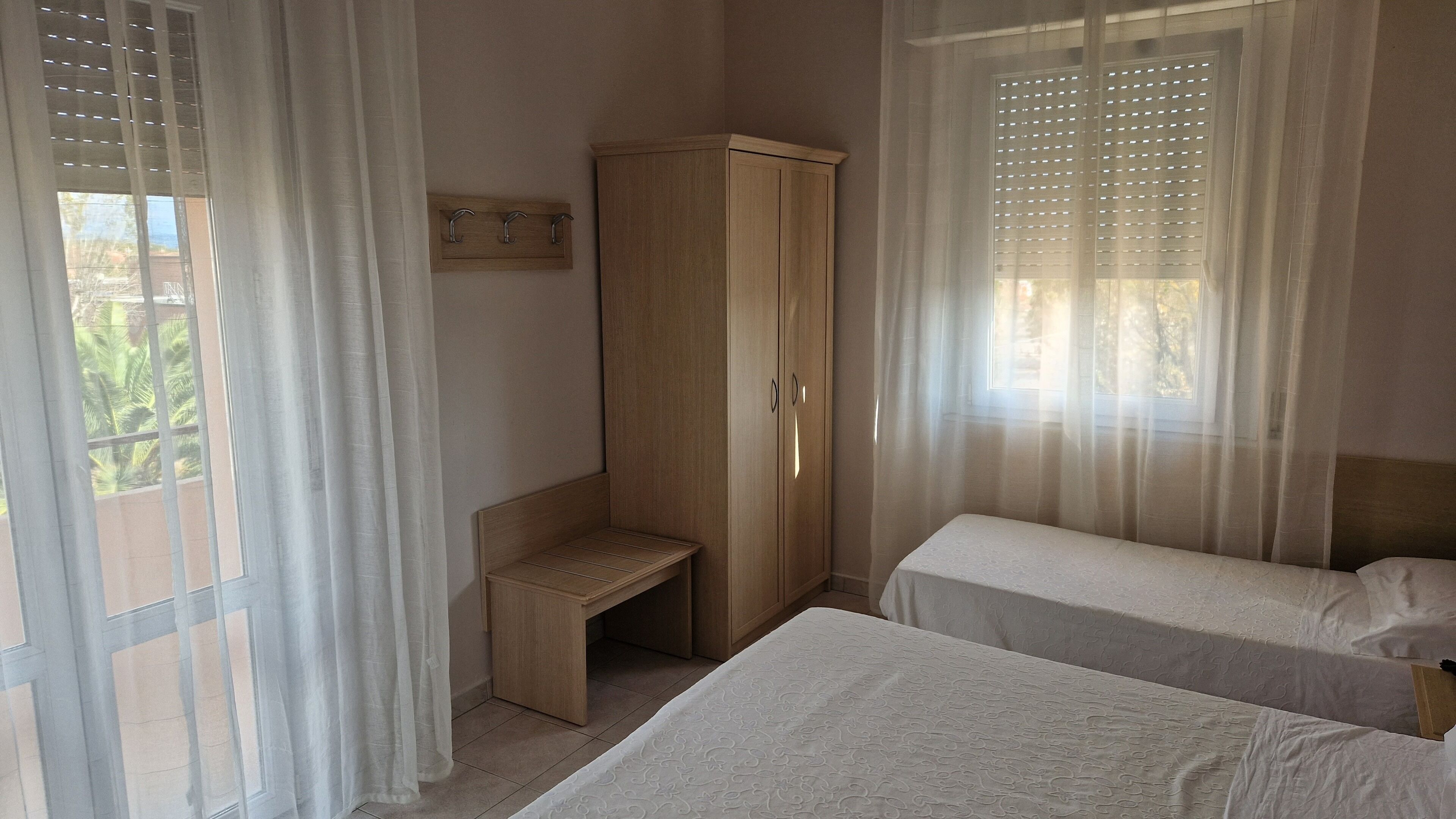 A bedroom with two beds, a wooden wardrobe, a bench, and a window with blinds.