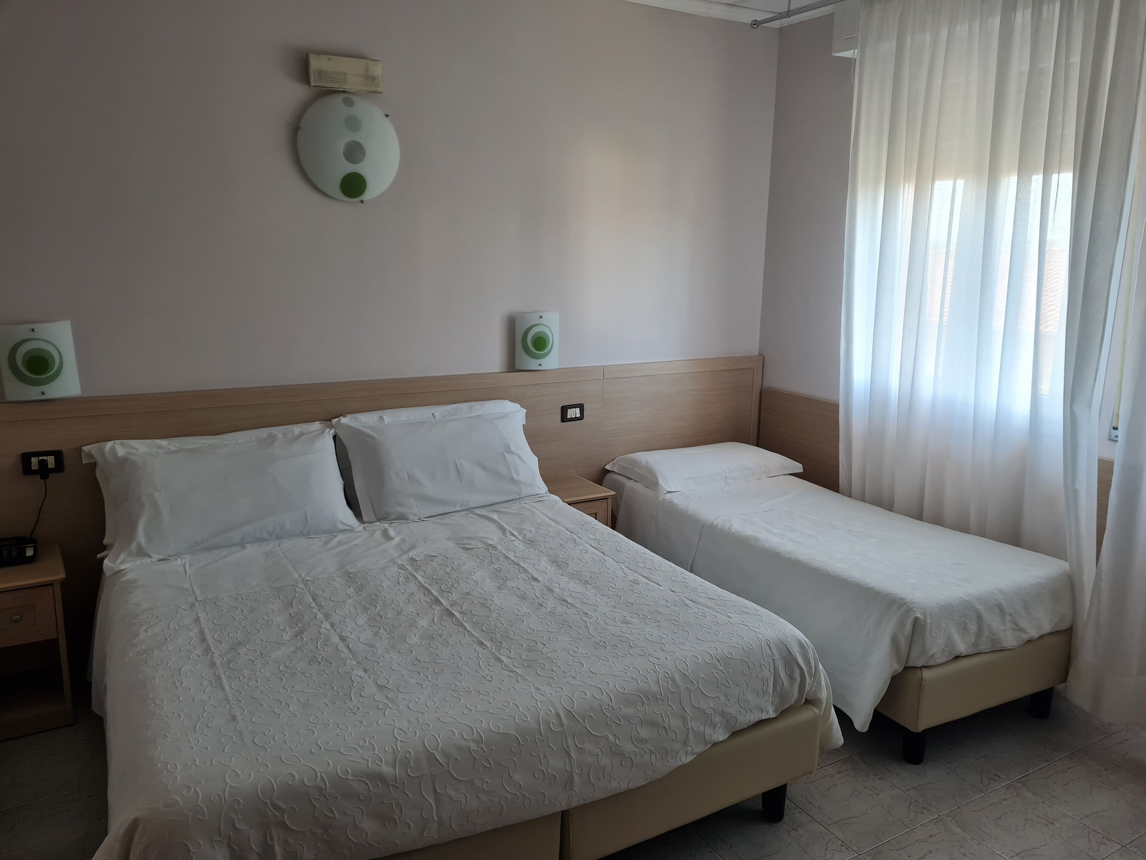 A hotel room with two beds, a wooden headboard, a bedside table with a lamp, and a window with sheer curtains.