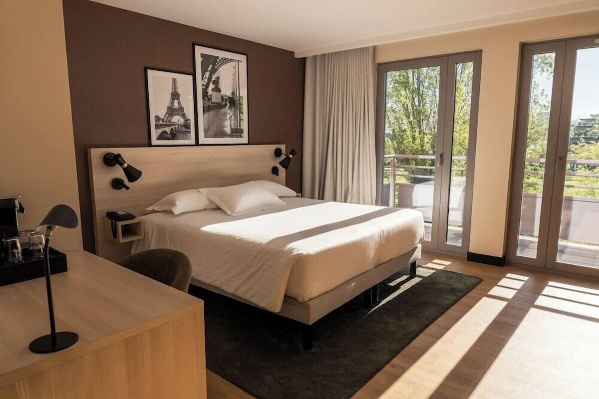 Een hotelkamer met een bed, een bureau, een stoel, een nachtkastje en een groot raam met uitzicht.