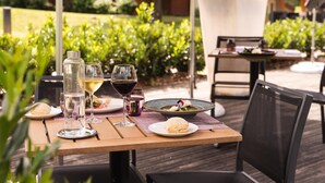 Un espace de restauration en plein air avec une table dressée pour deux personnes, comprenant une bouteille d’eau, des verres à vin et une assiette de nourriture.