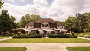 Un grand bâtiment à la façade symétrique, doté d’un espace extérieur aménagé avec des parasols et d’un jardin bien entretenu.